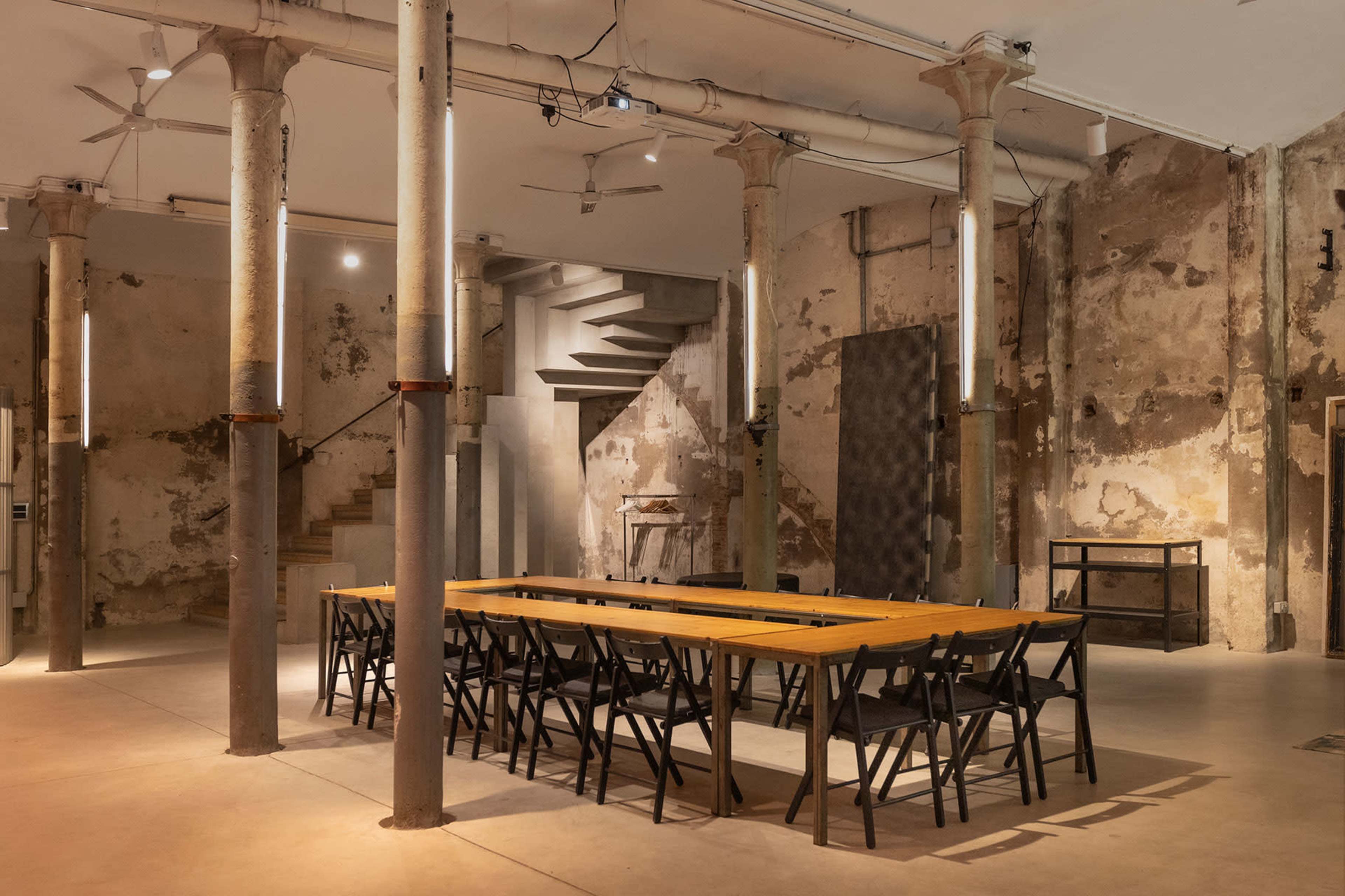 The image features a sparse, industrial-style room with a long wooden table surrounded by black chairs and tall columns, set against a backdrop of rough, partially bare walls.