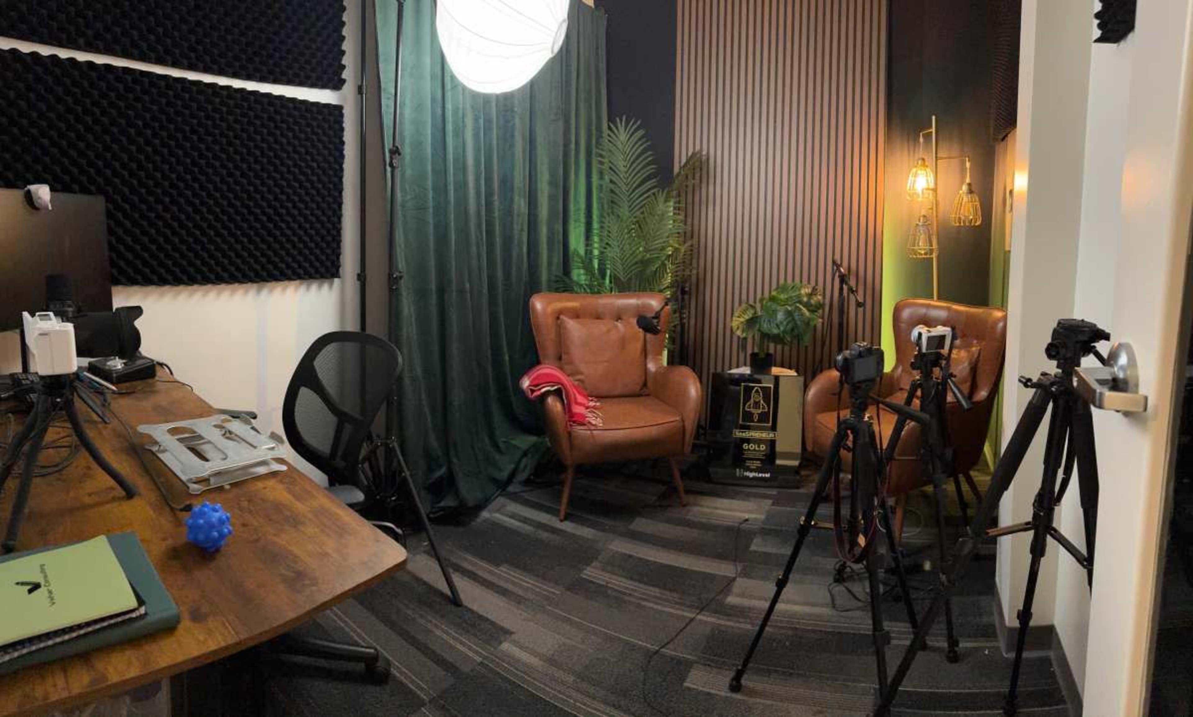 The image shows a small studio setup featuring two leather chairs, a wooden table with equipment, and several cameras positioned on tripods, surrounded by acoustic paneling and green curtains.