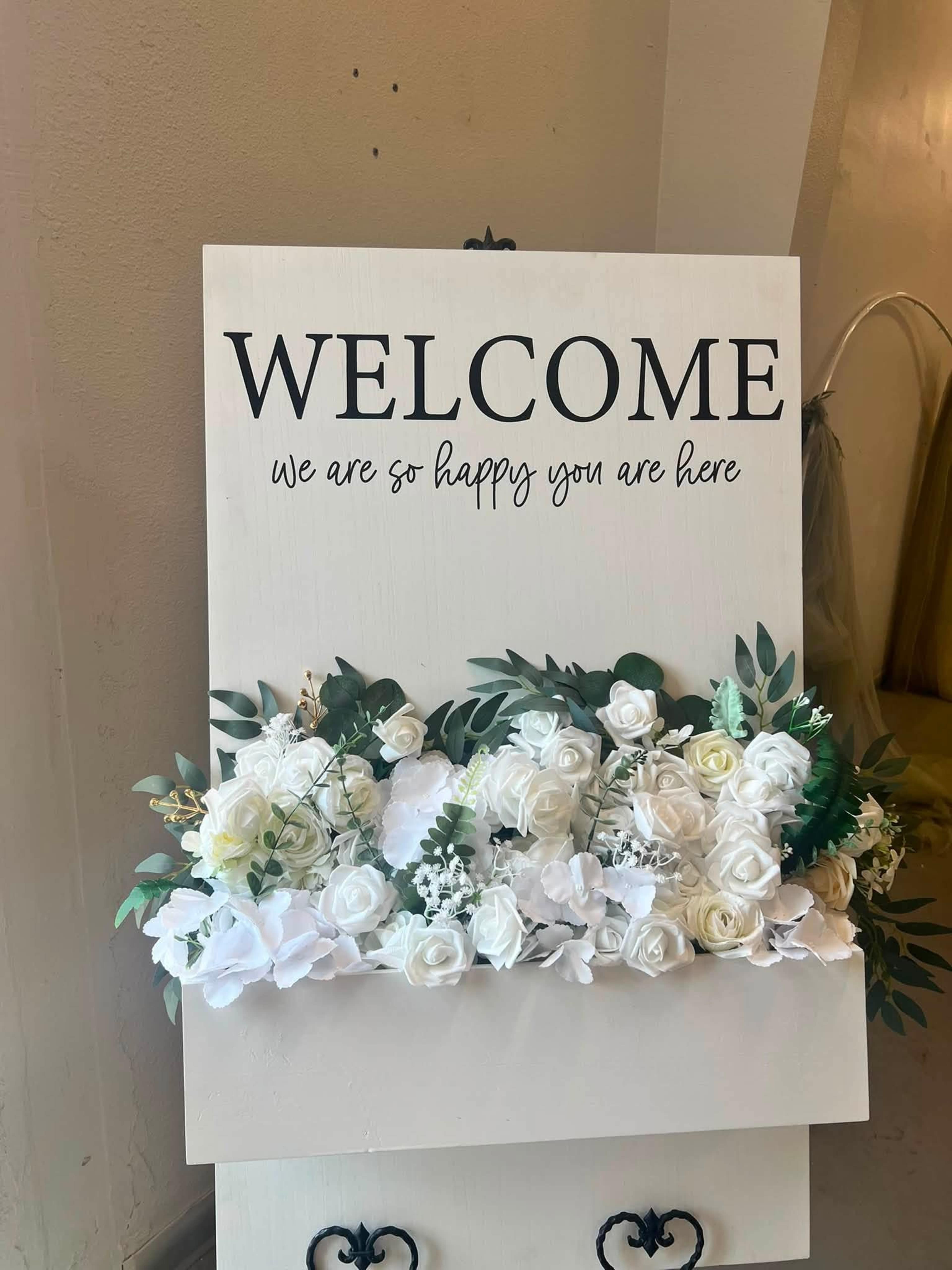 A welcome sign with the text "WELCOME" and "we are so happy you are here," adorned with a floral arrangement at its base, is displayed on a wall.