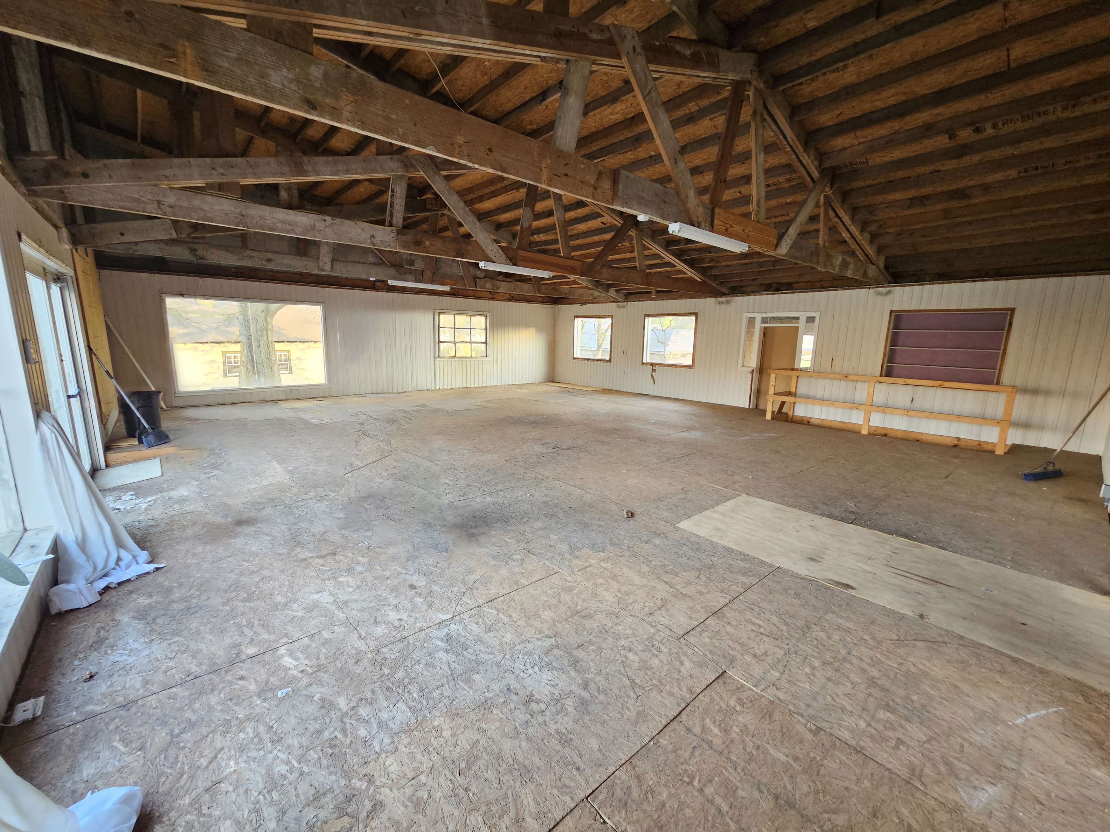 The image shows a spacious, empty room with wooden beams and large windows, featuring a bare floor and minimal furnishings.