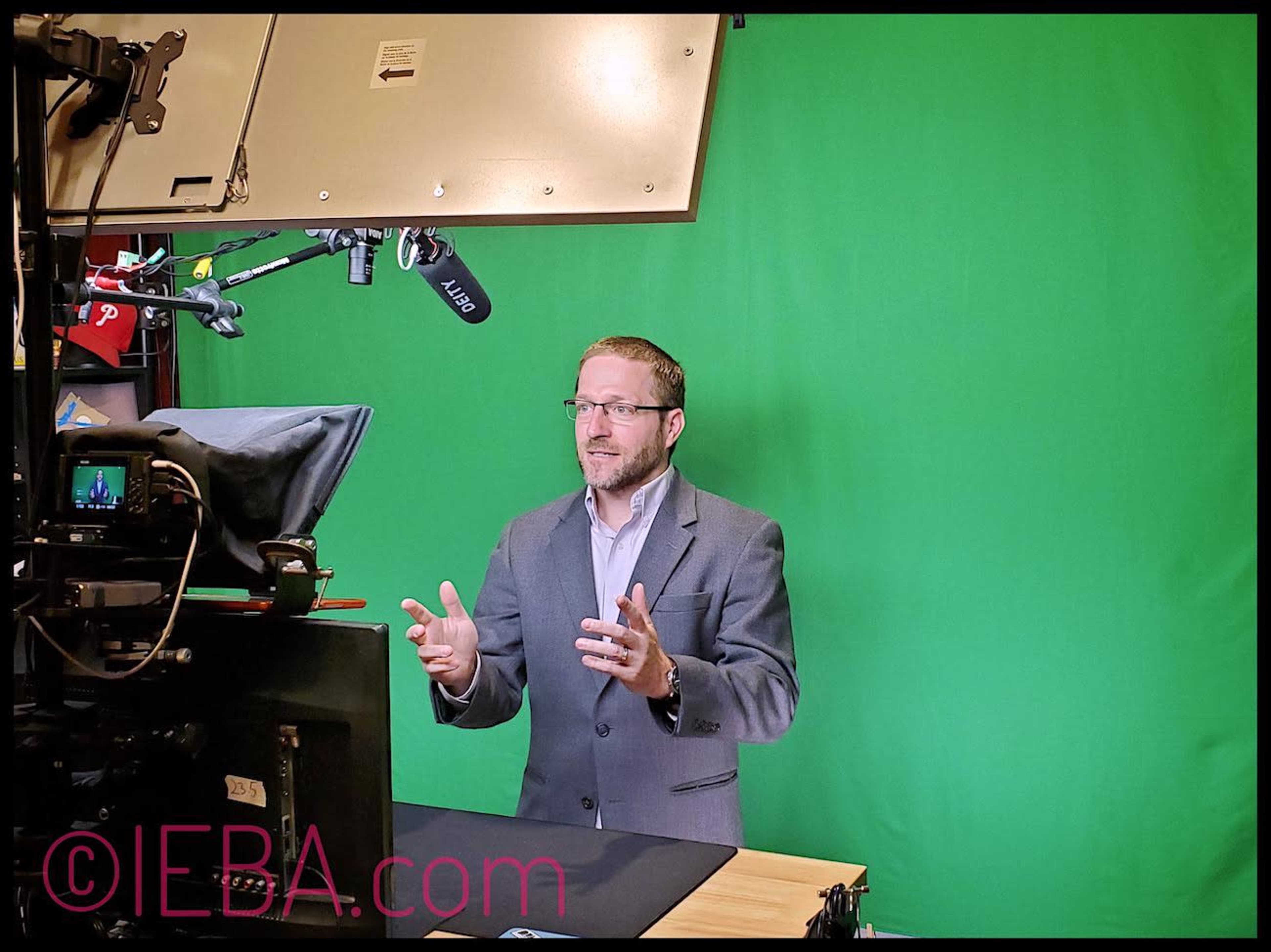 A man in a gray suit stands in front of a green screen while gesturing and speaking into a microphone.