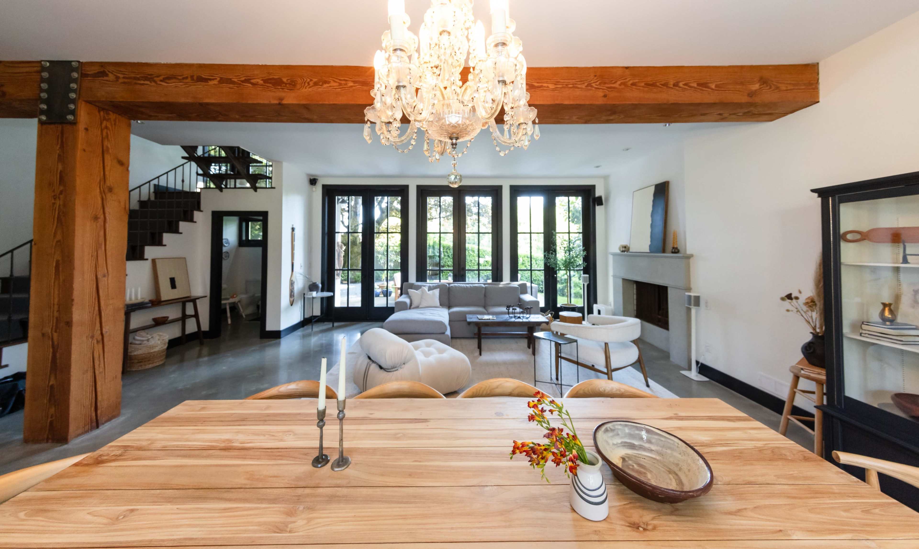 A spacious dining area with a wooden table and chandelier overlooks a living room featuring large windows and modern furnishings.