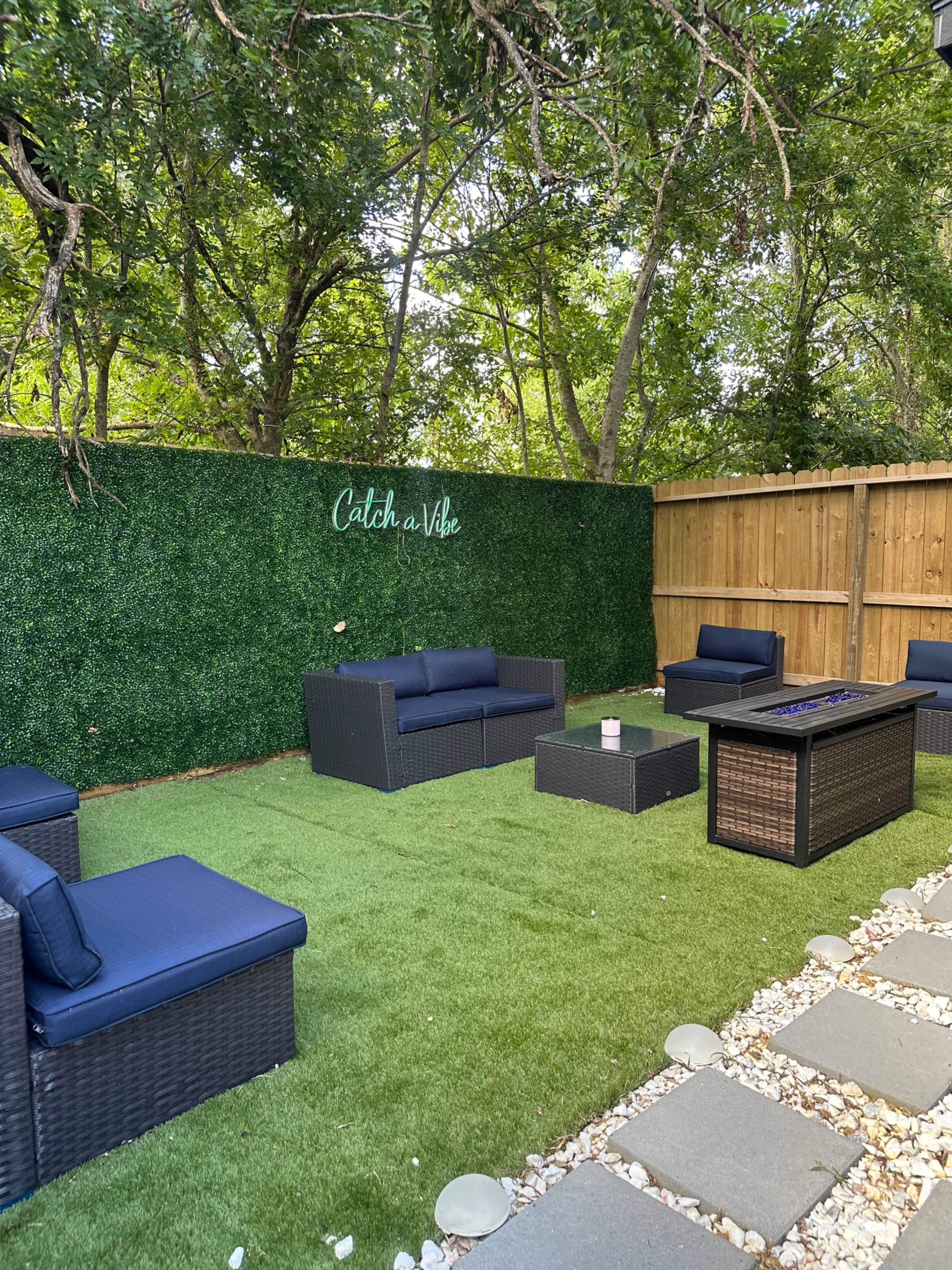 A outdoor seating area with dark wicker furniture arranged on a green artificial lawn, surrounded by trees and a wooden fence.