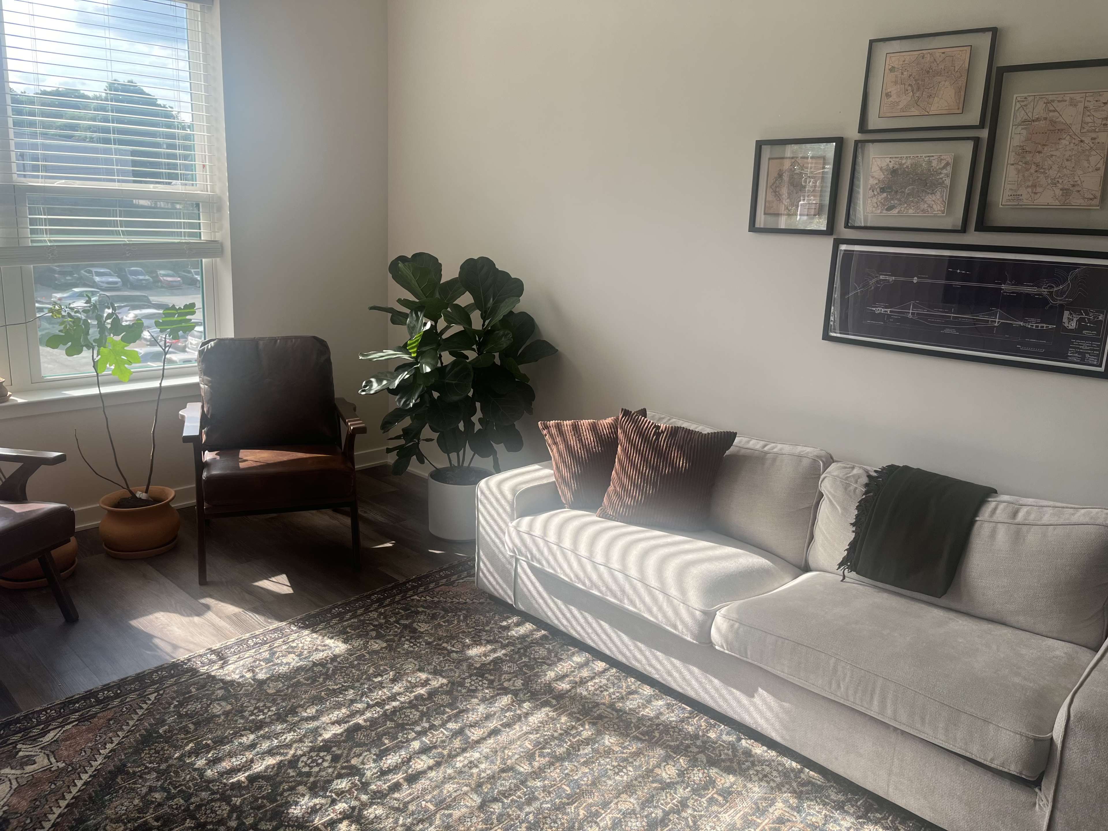 The image shows a well-lit living room featuring a gray sofa, a brown armchair, a potted plant, and framed maps on the wall.