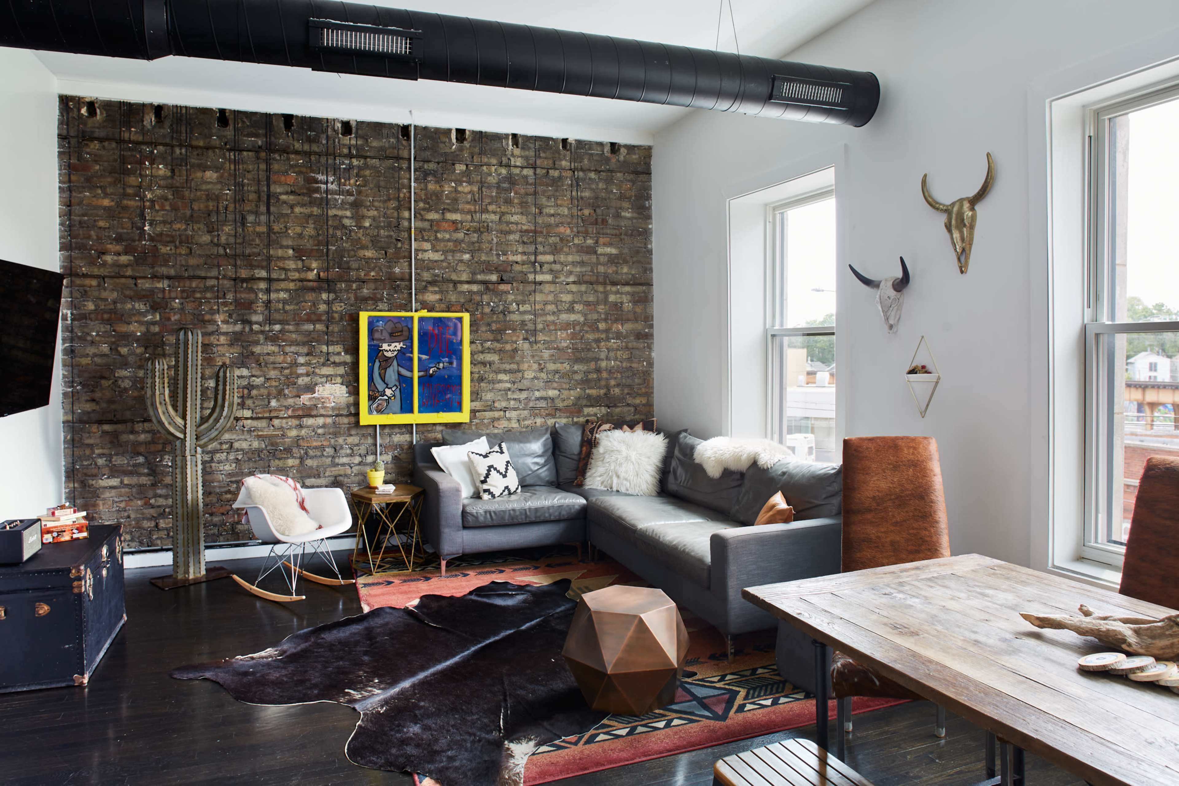 A modern living room with exposed brick walls, a sectional sofa, a coffee table, decorative art, and large windows providing natural light.