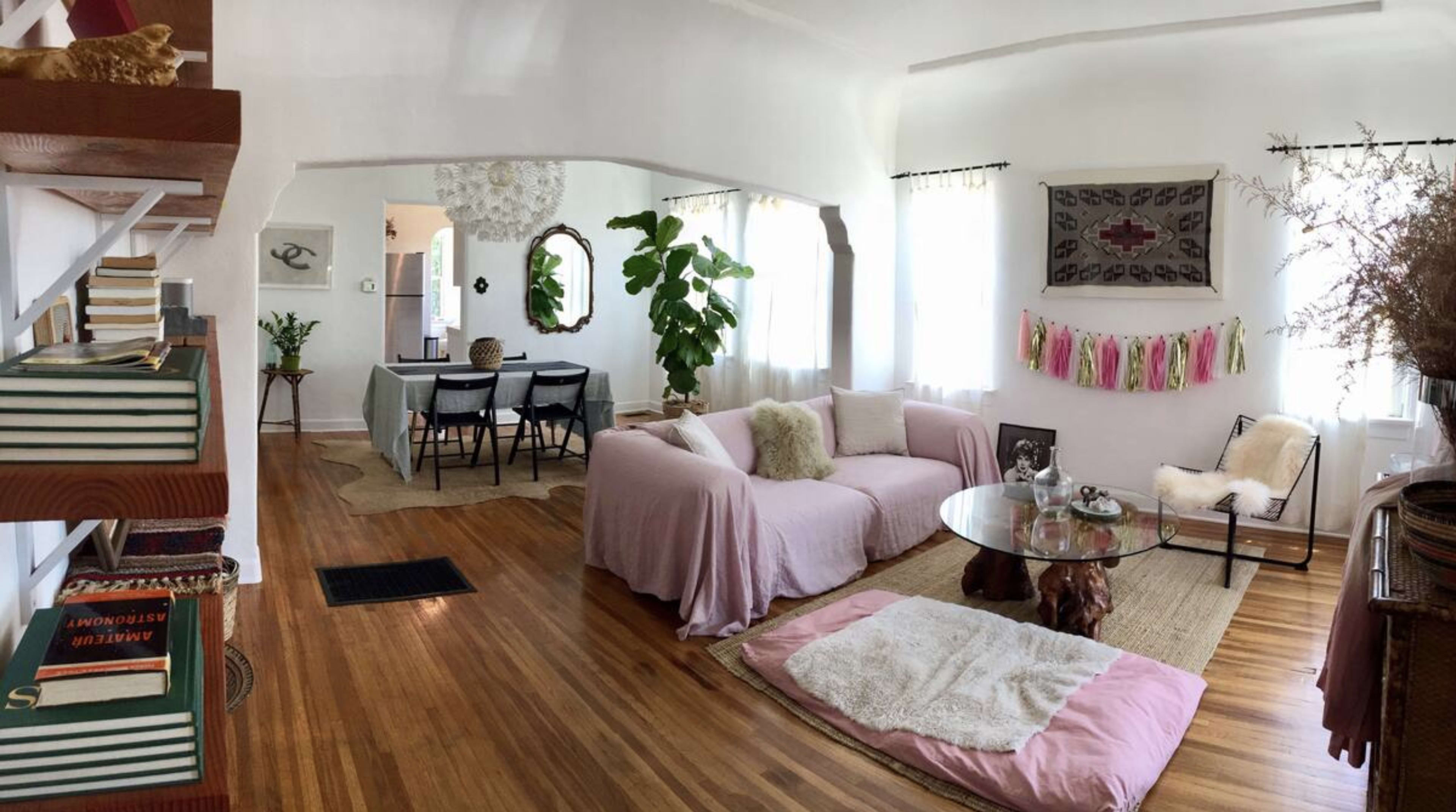 The image shows a living room with a pink sofa, wooden floor, a glass coffee table, and a dining area visible in the background.