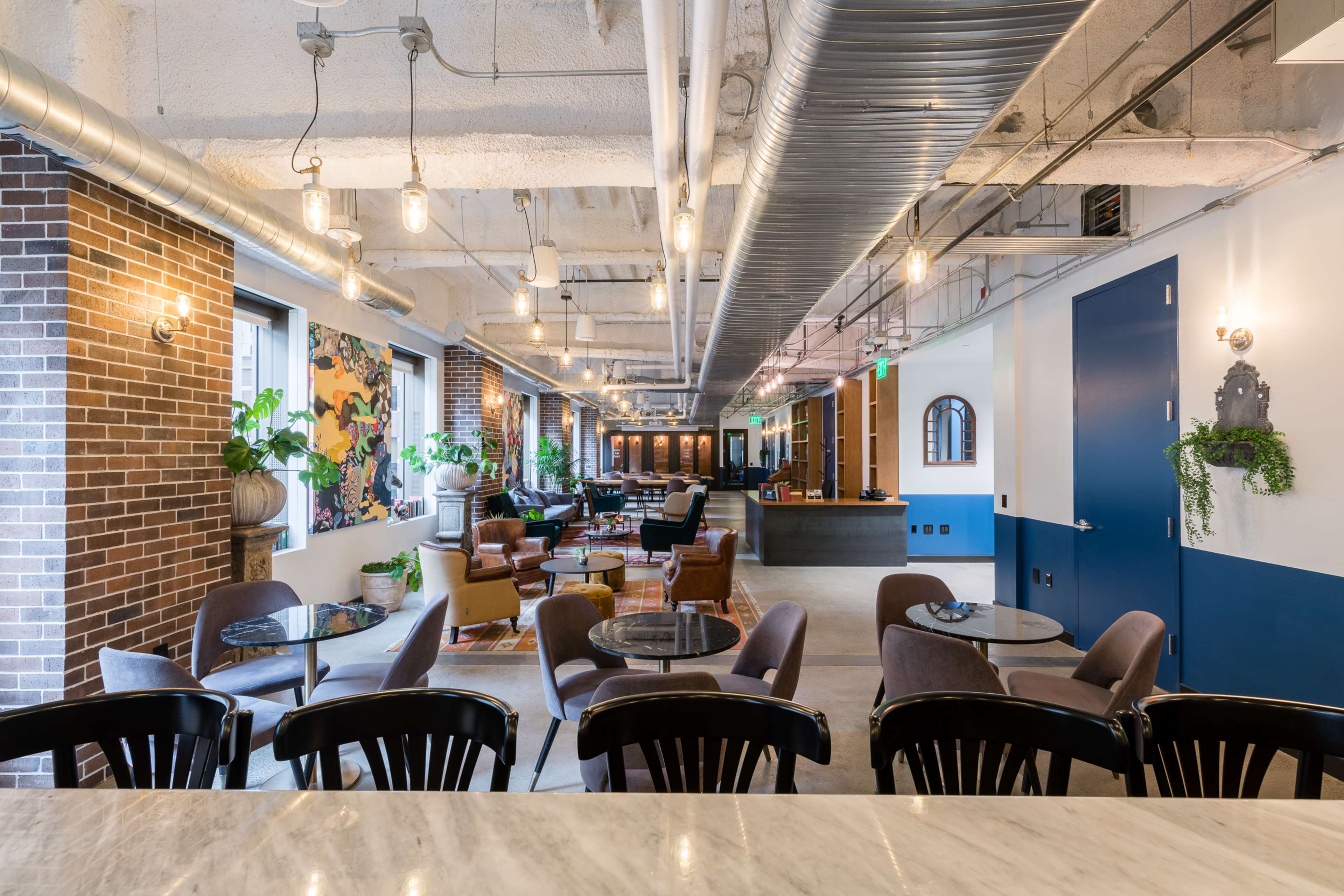 A modern, spacious coworking space featuring seating areas, tables, and decorative plants, with exposed brick walls and industrial lighting.