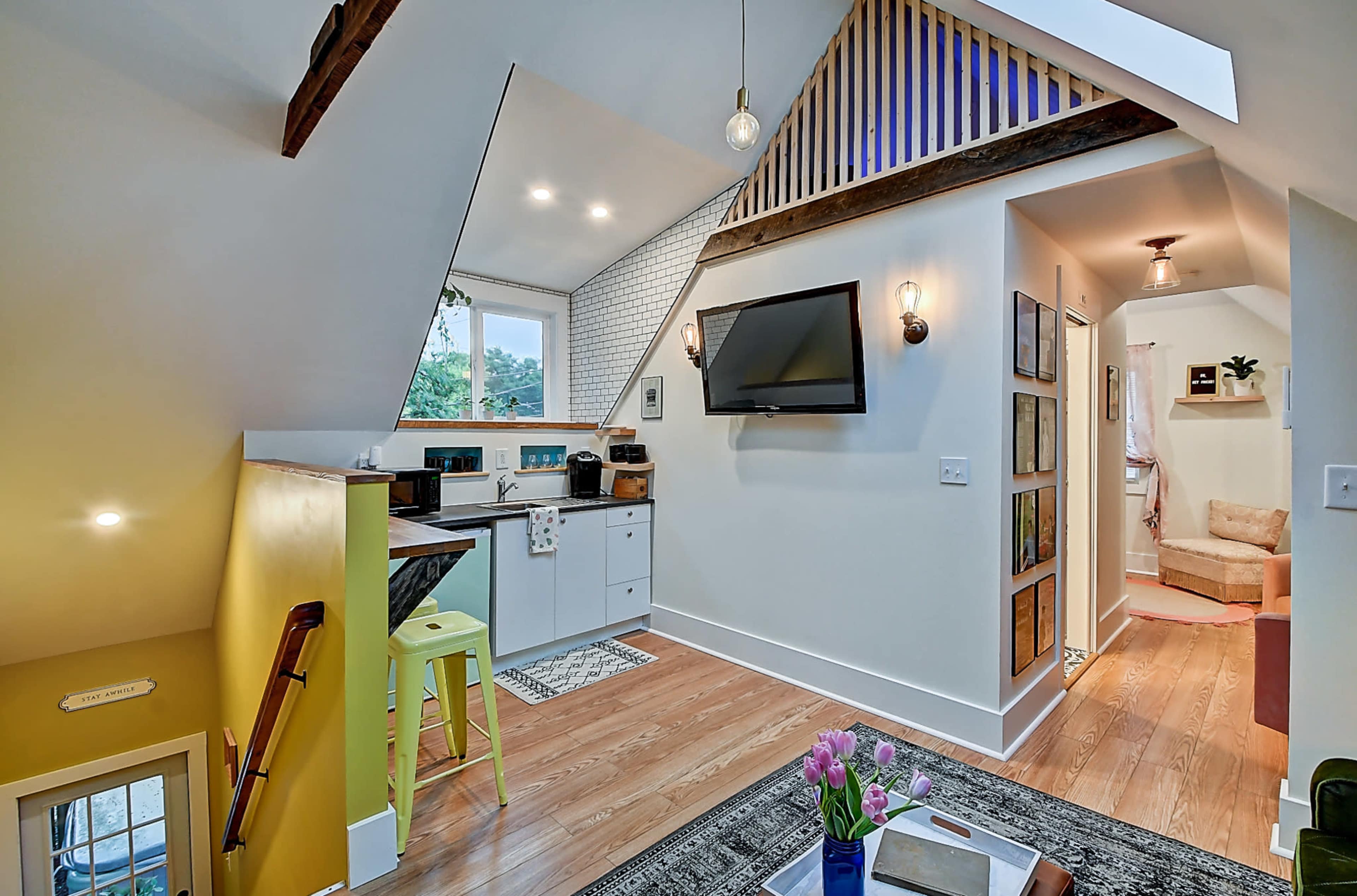 The image shows a modern, cozy interior of a small house featuring a kitchenette, a living area with a television, and a staircase leading to an upper level.