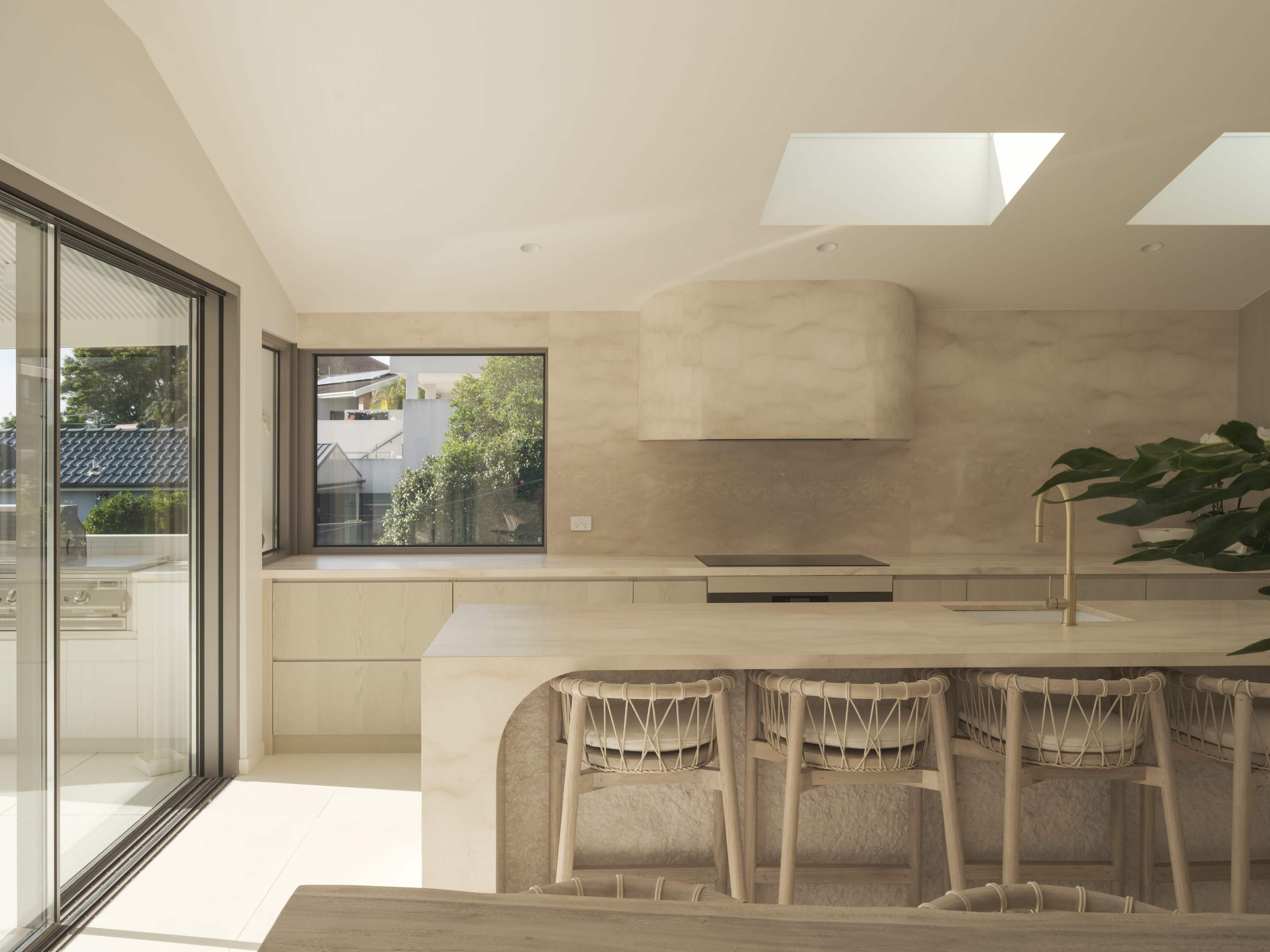 The image shows a modern kitchen with a large island, bar seating, and large windows allowing natural light to enter.