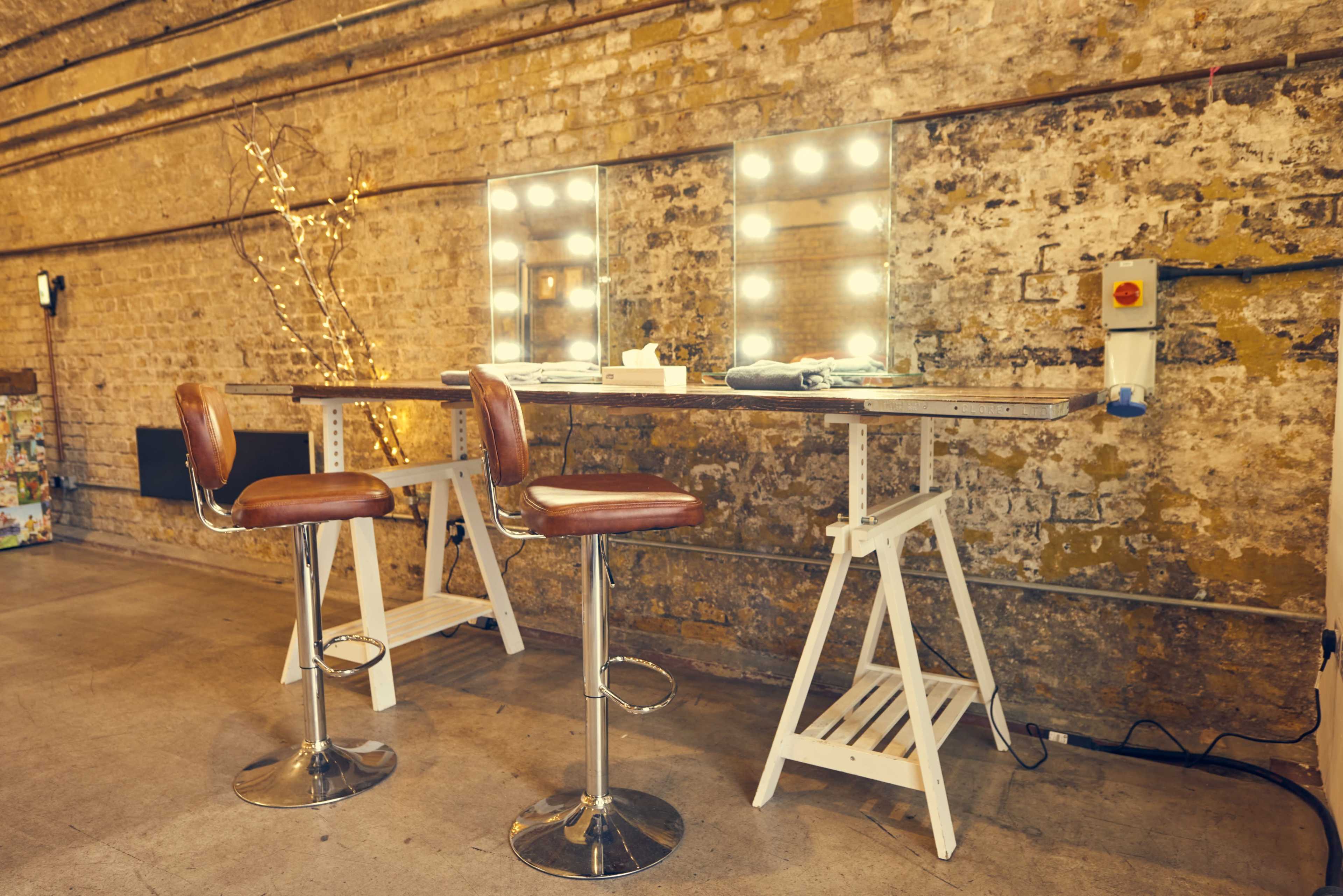 A makeup station with two high stools, a wooden table, and three illuminated mirrors is set against a brick wall.
