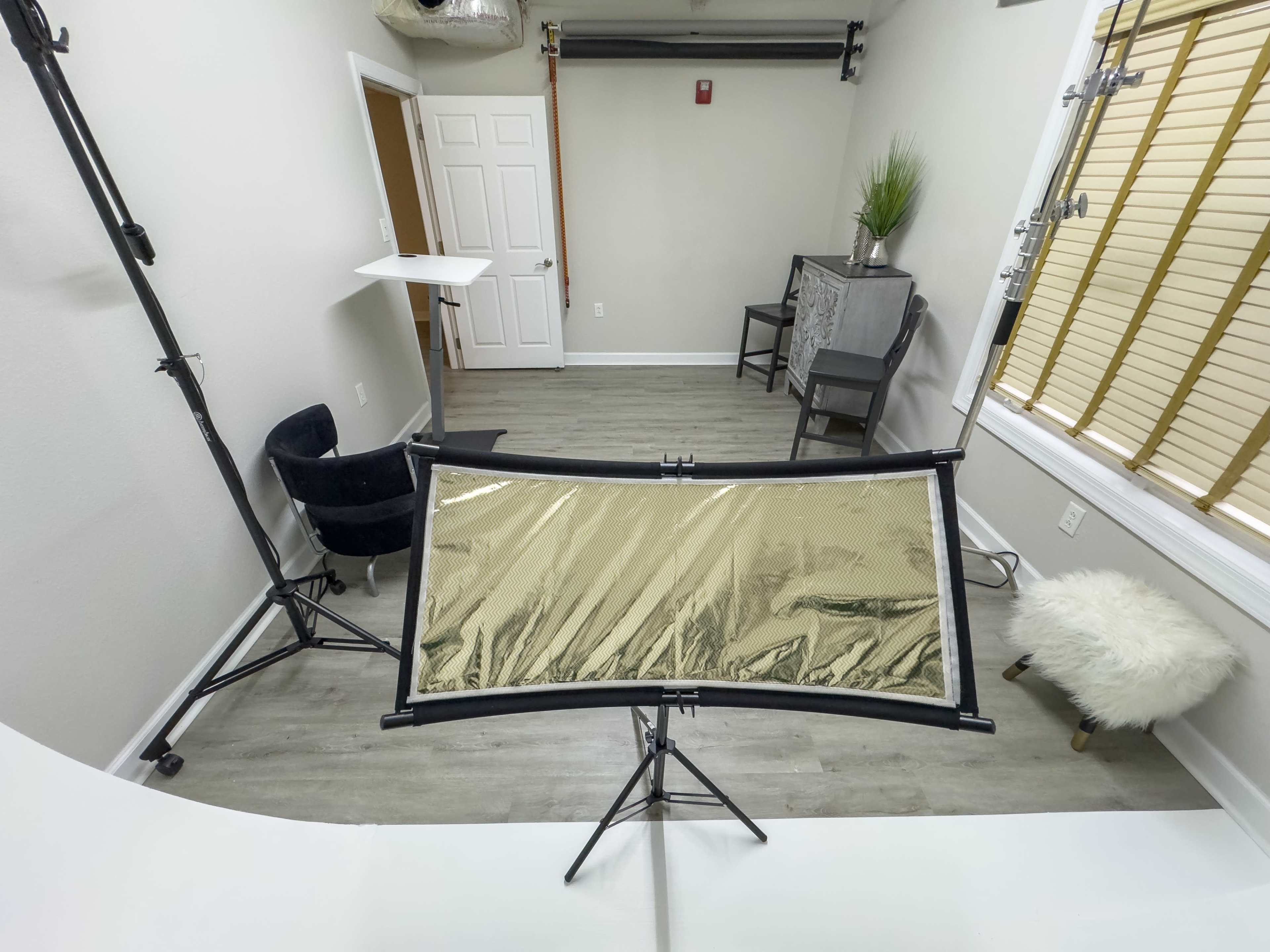 A well-lit studio setup featuring a large reflector on a stand, a chair, a small table, and a decorative plant against a wall with wooden blinds.