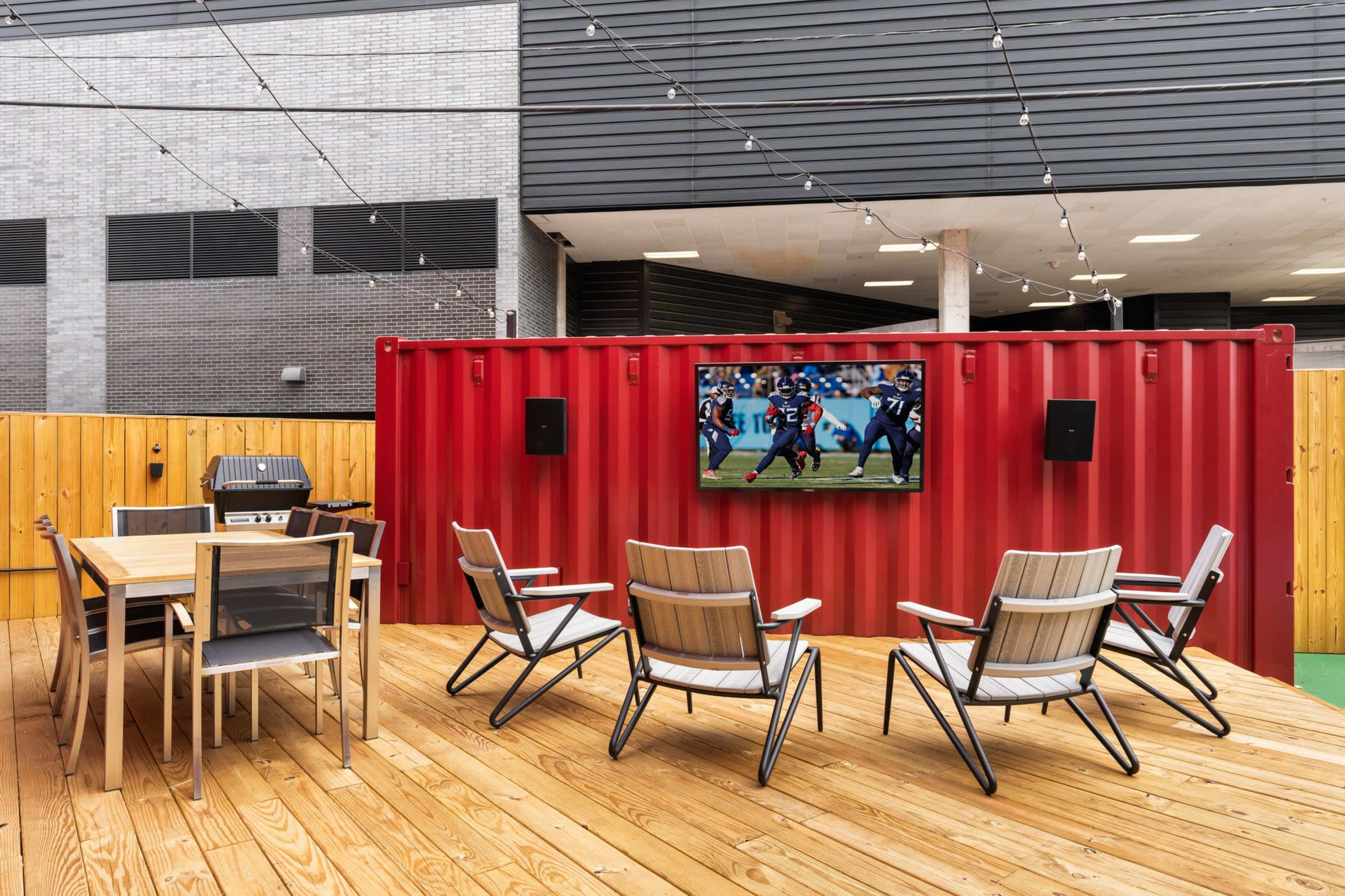 The image shows a modern outdoor seating area with a wooden deck, featuring a table and chairs, a grill, and a television mounted on a red shipping container wall.