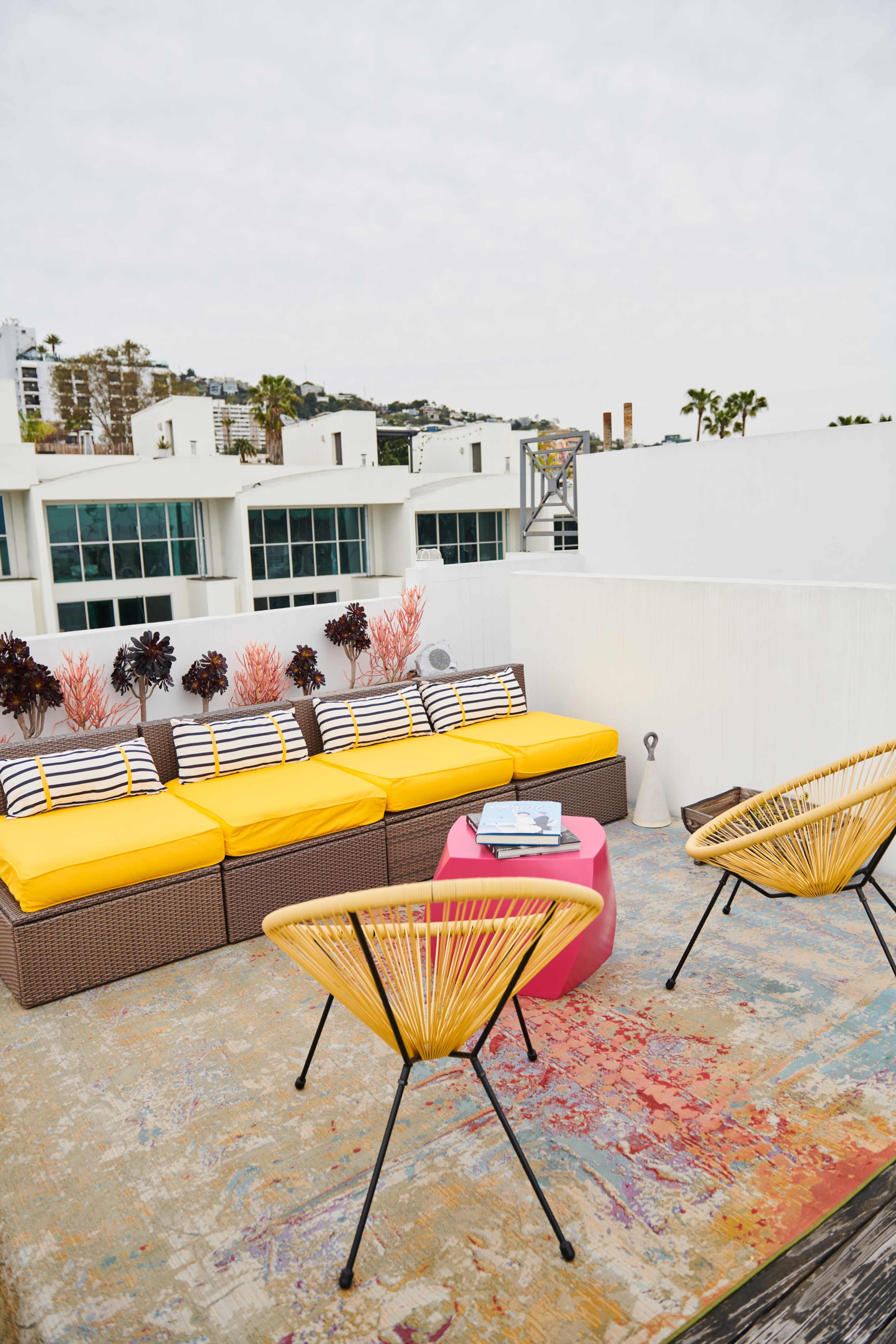 A rooftop seating area features a yellow sofa with striped cushions, two rattan chairs, and a pink side table surrounded by decorative plants and a view of buildings in the background.
