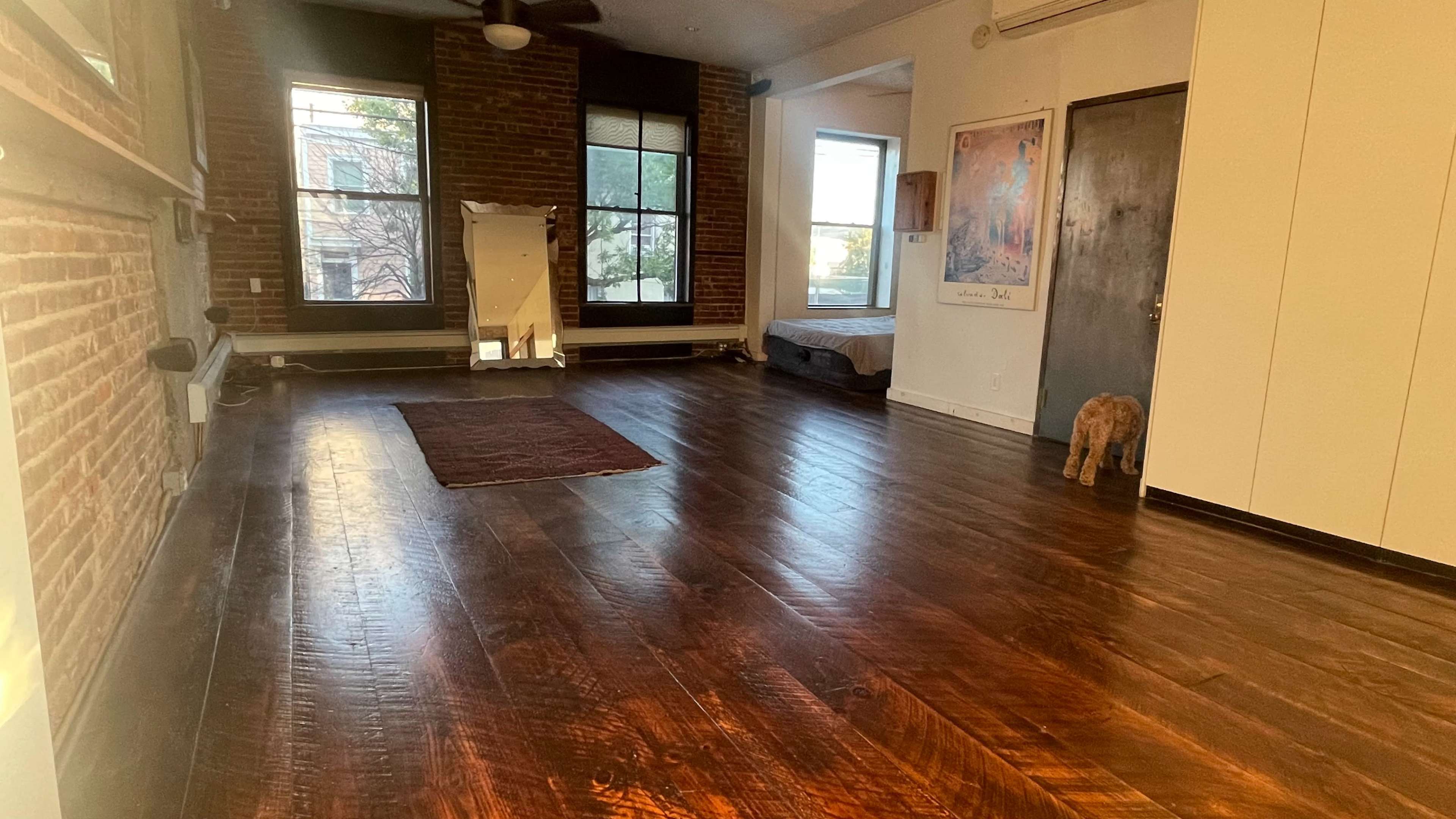 A spacious room featuring hardwood floors, brick walls, large windows, and a mirror, with a bed and a dog near the door.