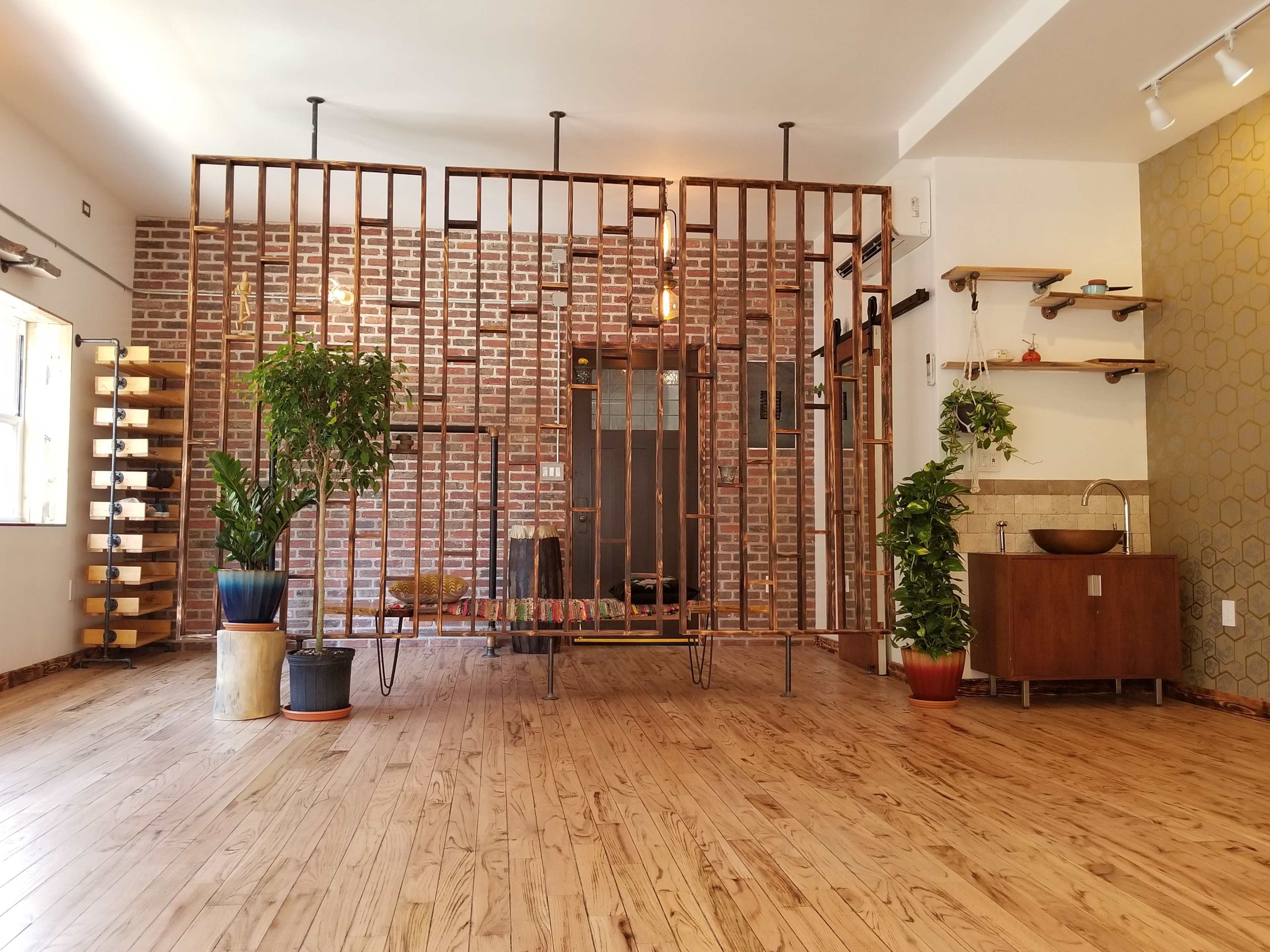A spacious room with wooden flooring, featuring a bamboo partition, potted plants, and a brick wall in the background.