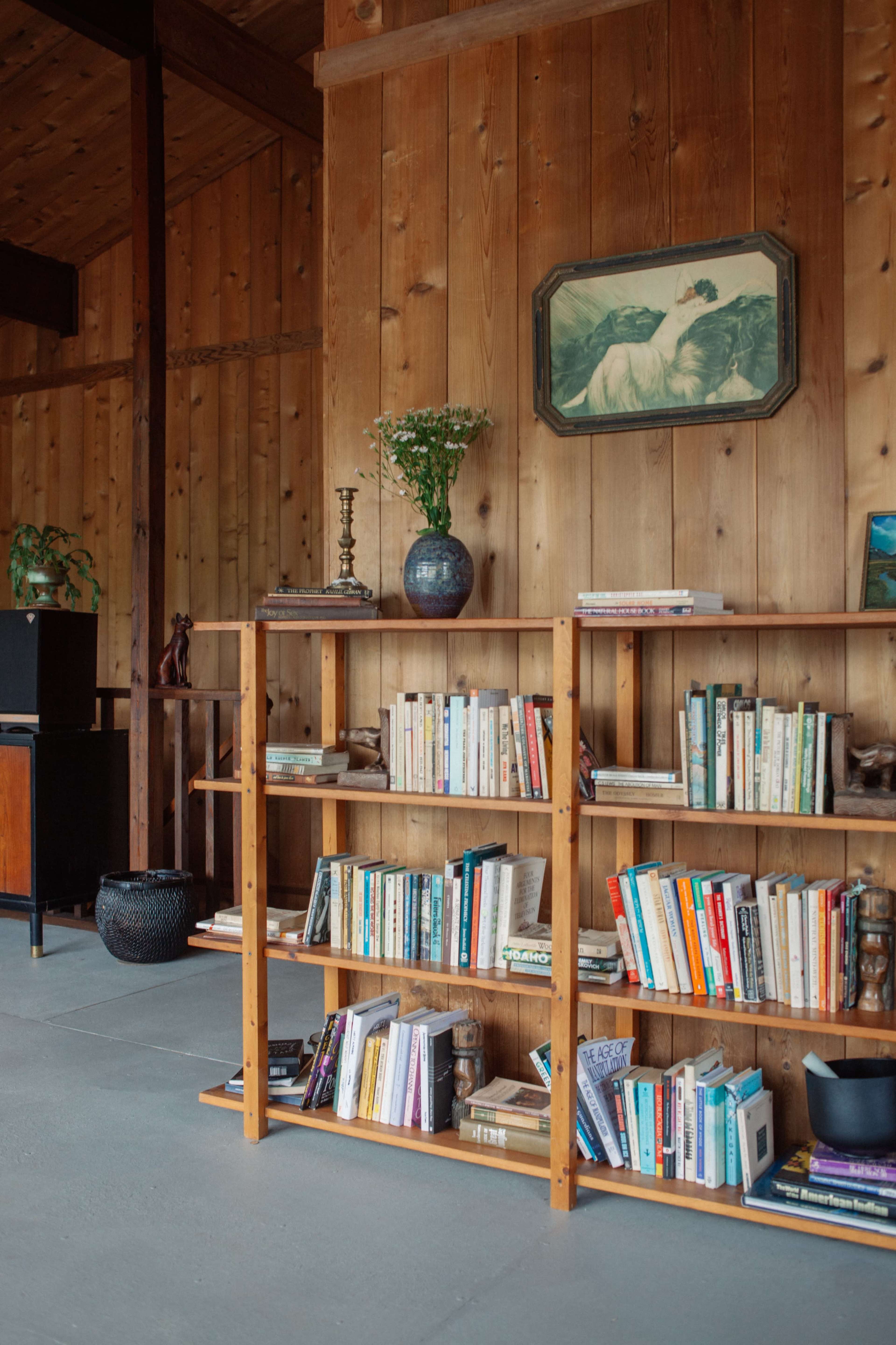 A wooden bookshelf filled with various books, positioned against a wall of wooden panels, with a framed painting and plants on display above it.