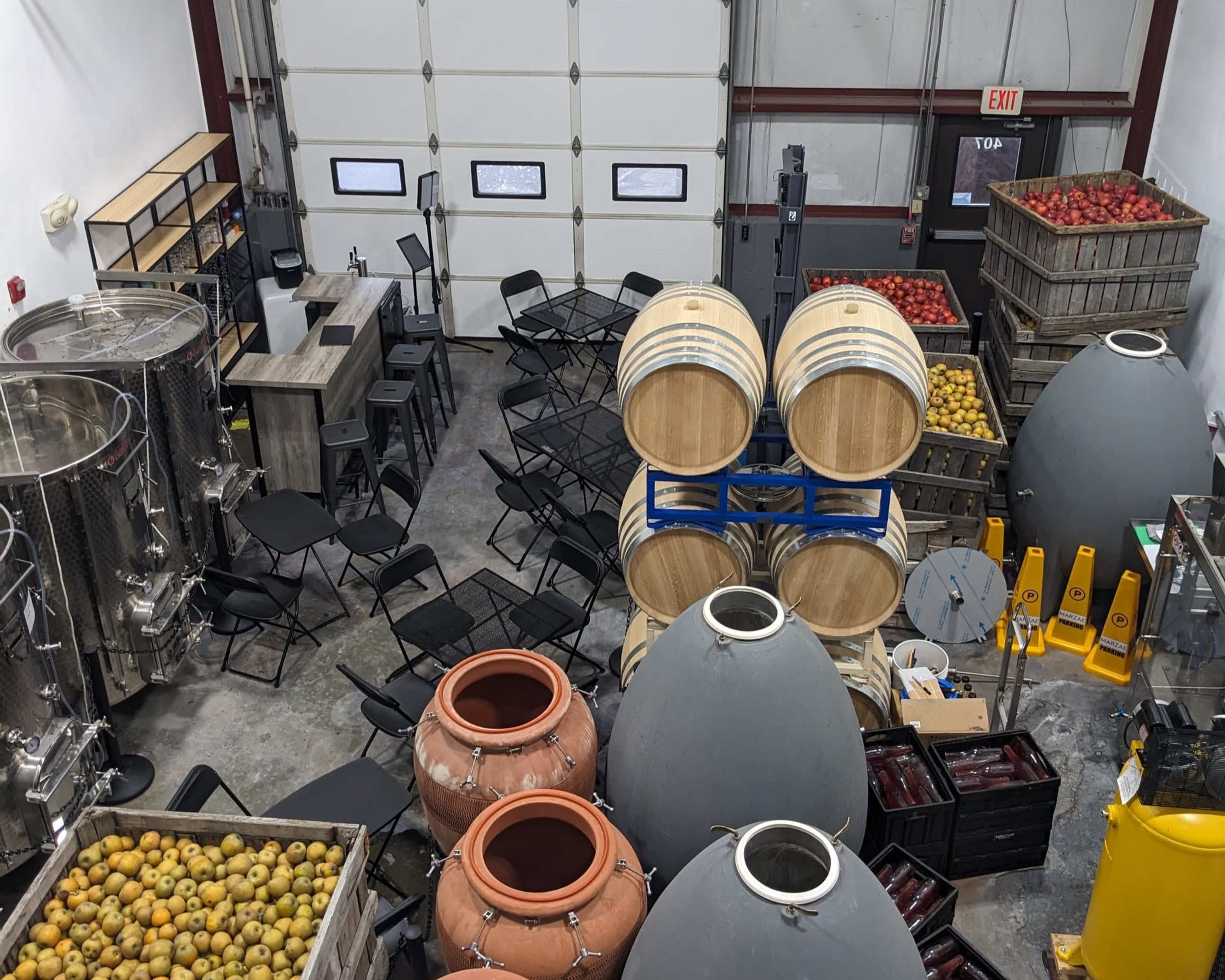 A winery interior with fermentation tanks, barrels, amphorae, tables, chairs, and crates of apples in an industrial-style space.