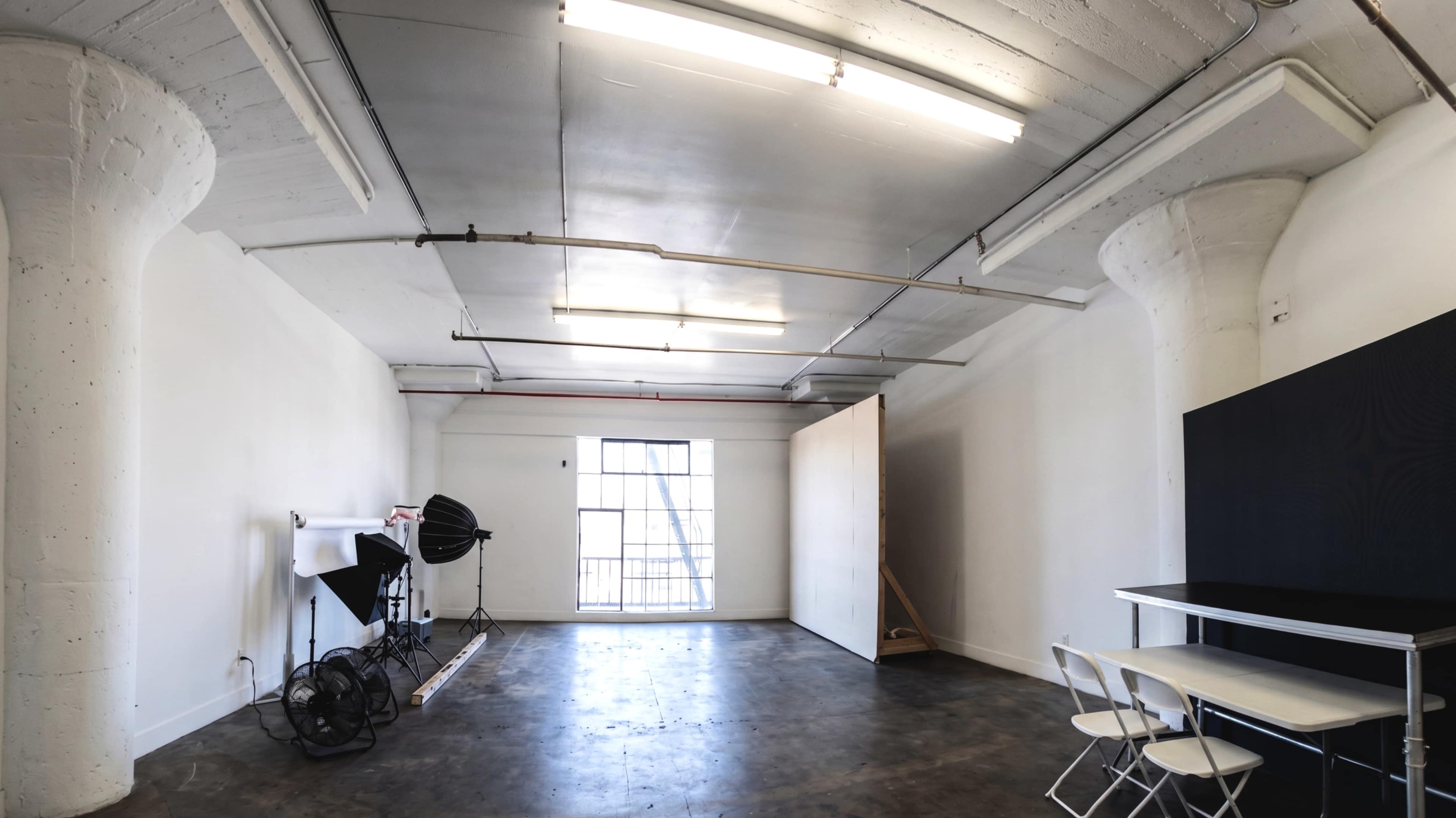 The image shows a spacious, empty studio featuring concrete pillars, a window allowing natural light, and minimalist furniture, including a table and chairs.