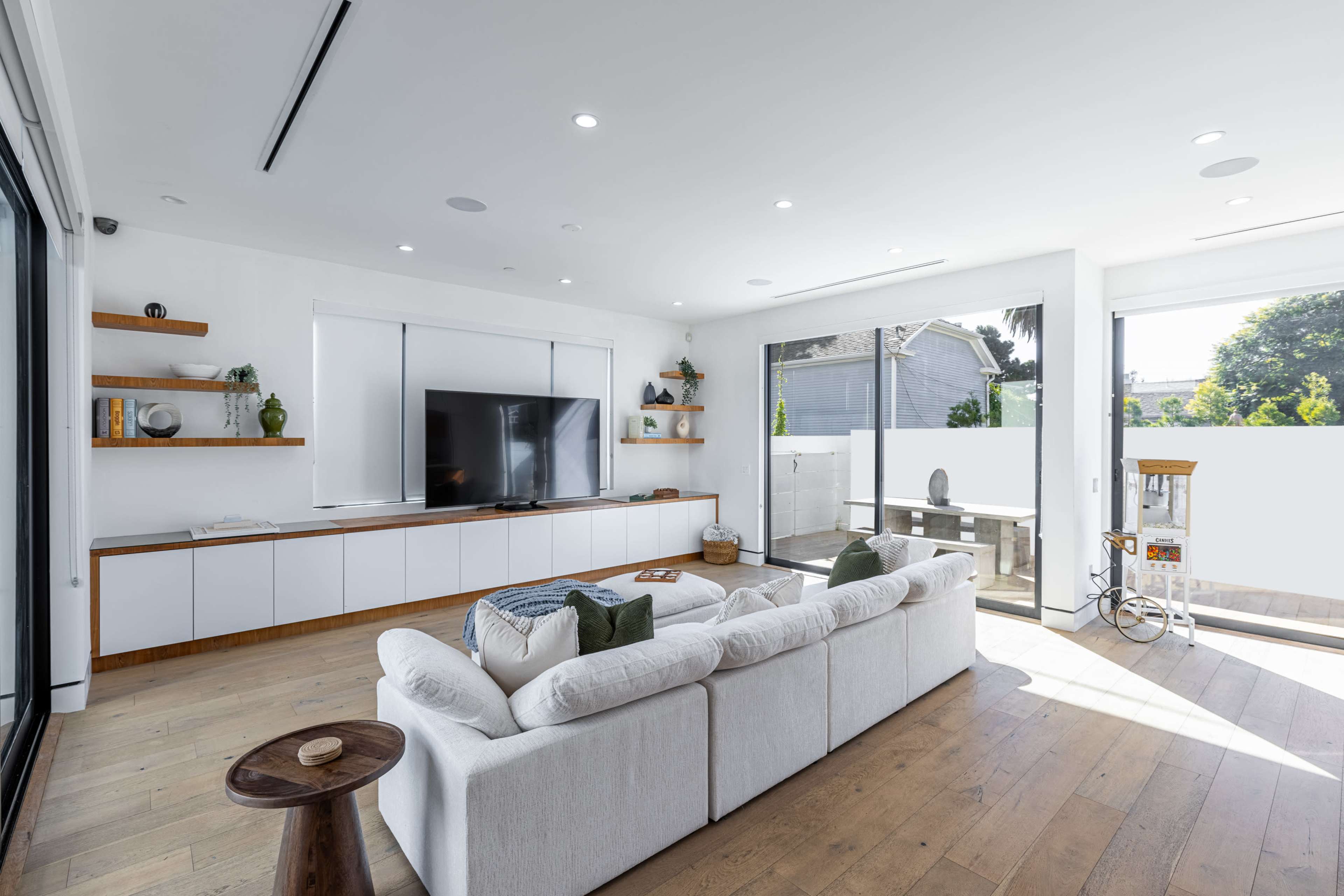 A modern, bright living room with a large TV mounted on the wall, a white sectional sofa, and sliding glass doors leading to an outdoor area.