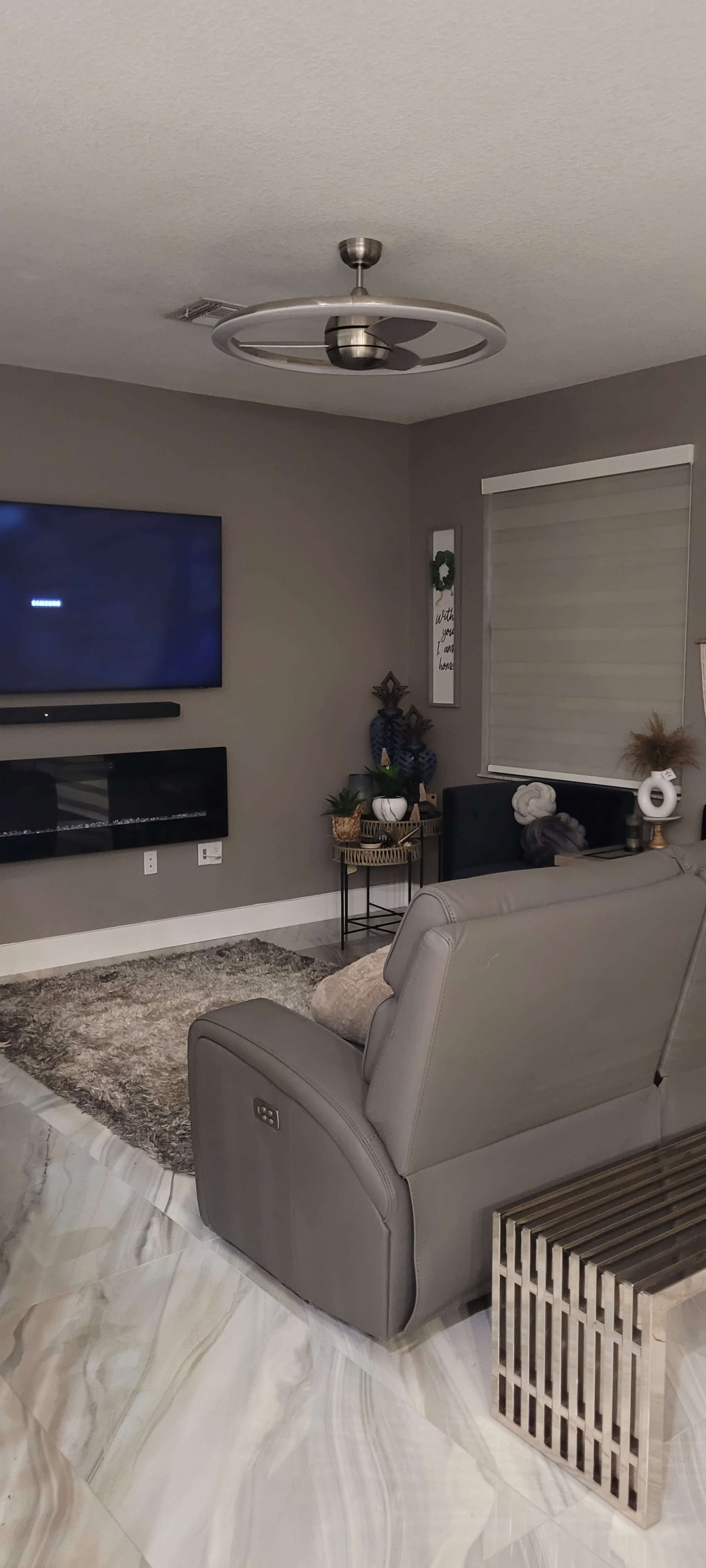 A modern living room with a large television mounted on the wall, a gray recliner, and decorative plants in a basket beside a fireplace unit.