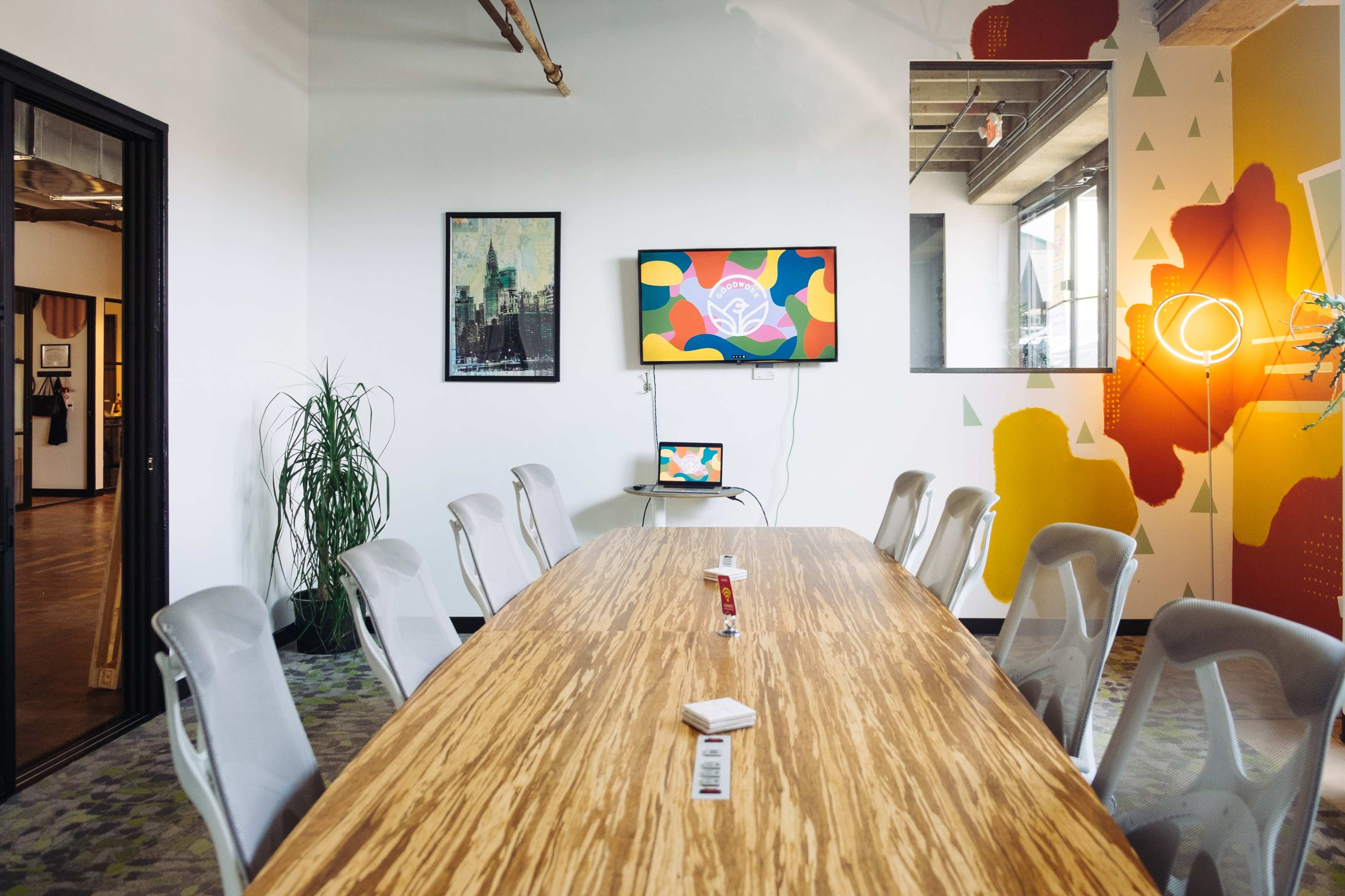 A modern conference room features a long wooden table, several ergonomic chairs, a wall-mounted TV displaying colorful artwork, and vibrant wall decorations.