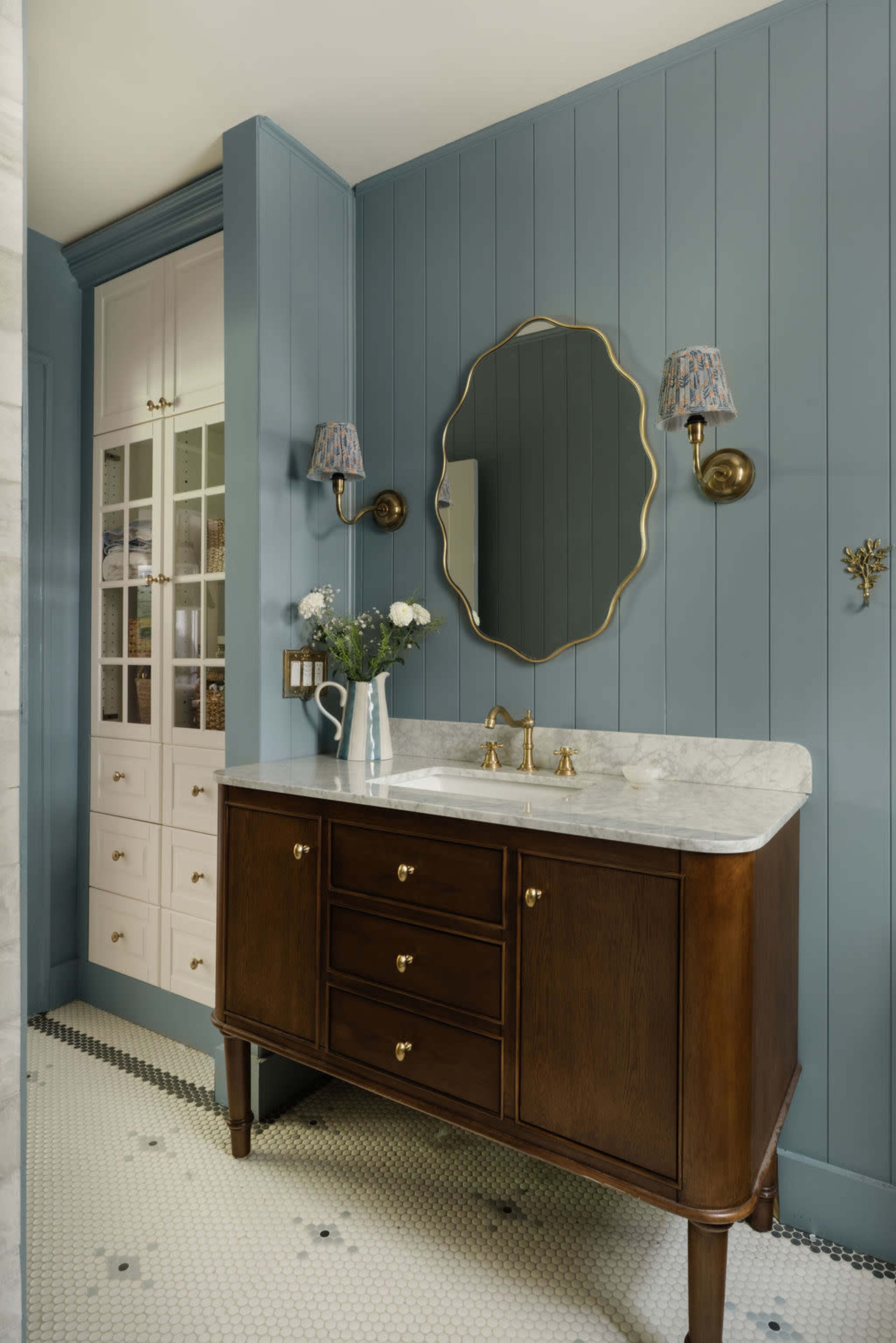 The image shows a bathroom featuring a wooden vanity with a marble countertop, a round mirror, and blue paneled walls, accompanied by wall sconces and decorative elements.