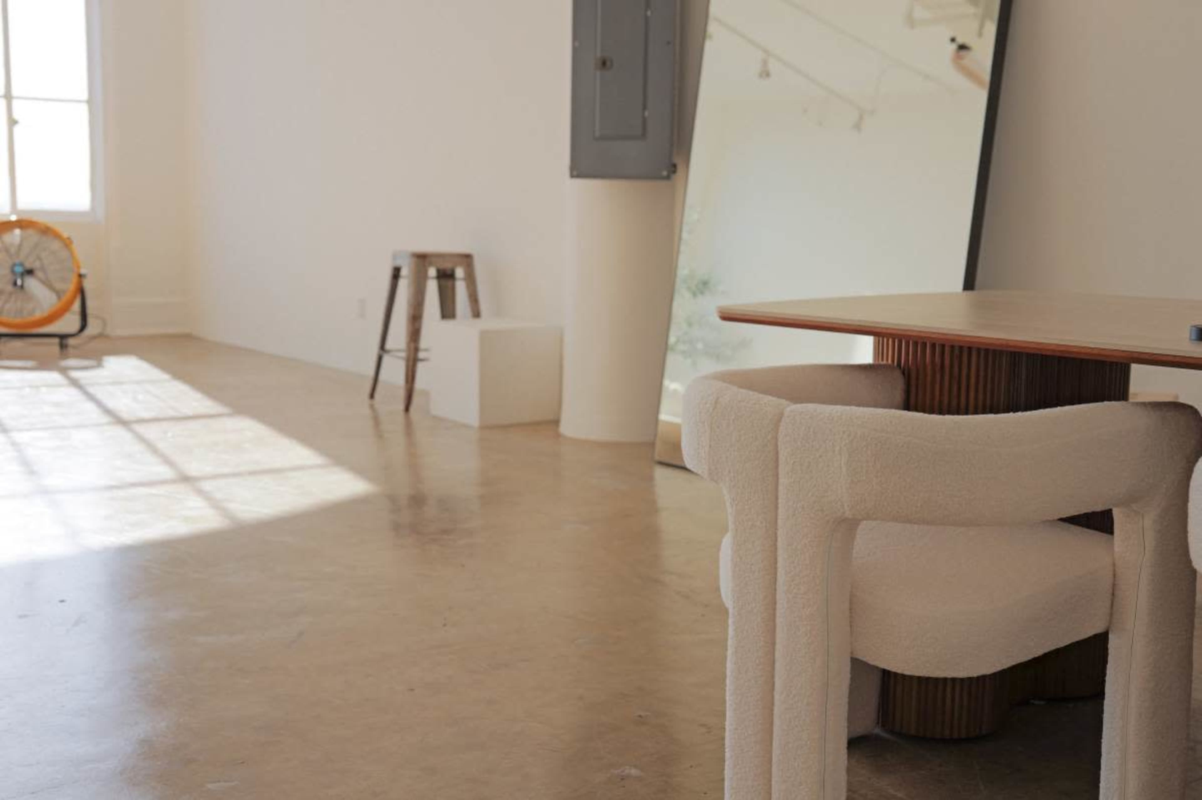 The image shows a modern, minimalist interior featuring a light-colored floor, a wooden table with a secured seating area, and a large mirror leaning against a wall.