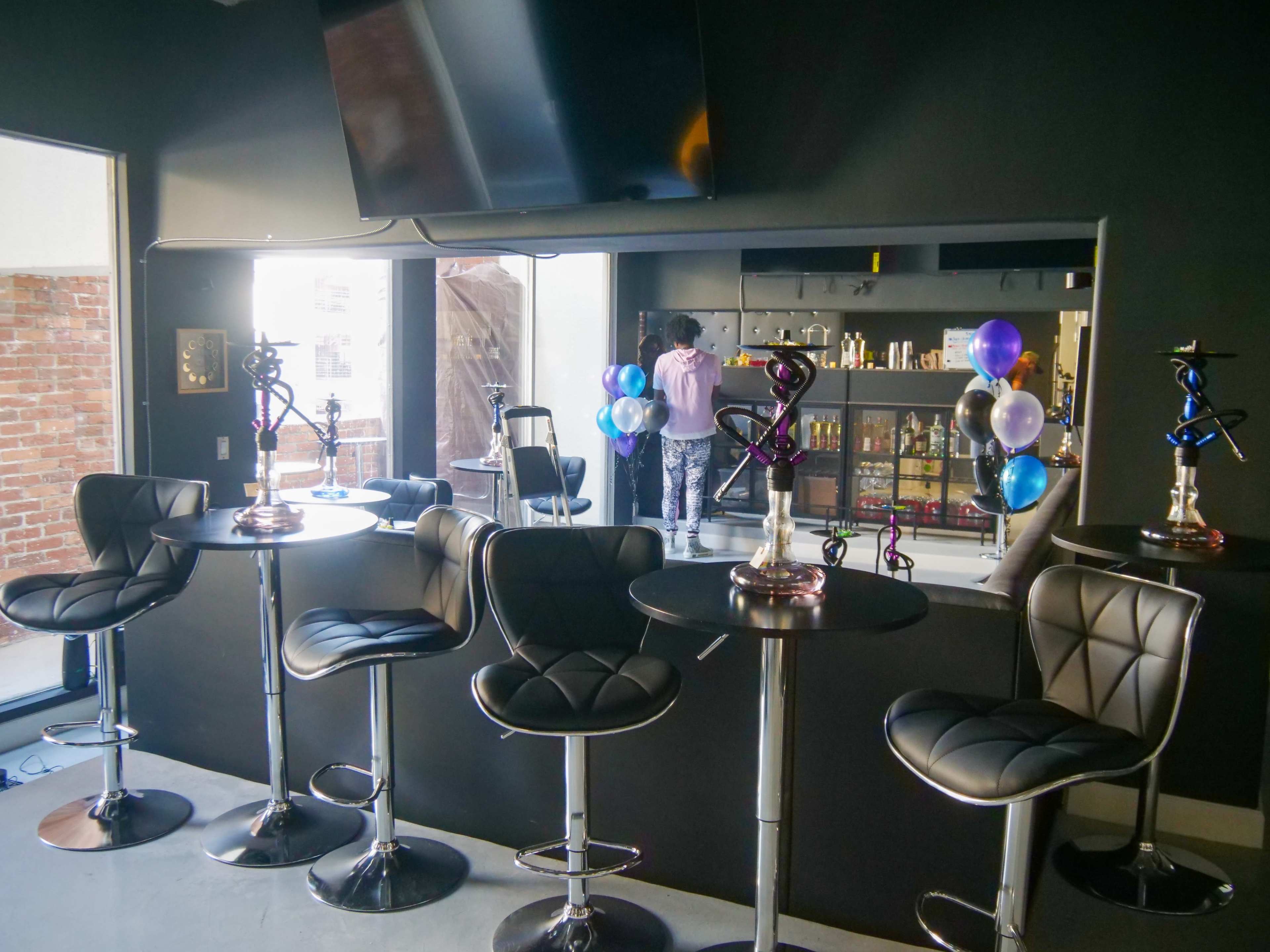 A modern bar setup features high-top tables and stools, with a black wall and a TV, while a person adjusts decorations nearby.