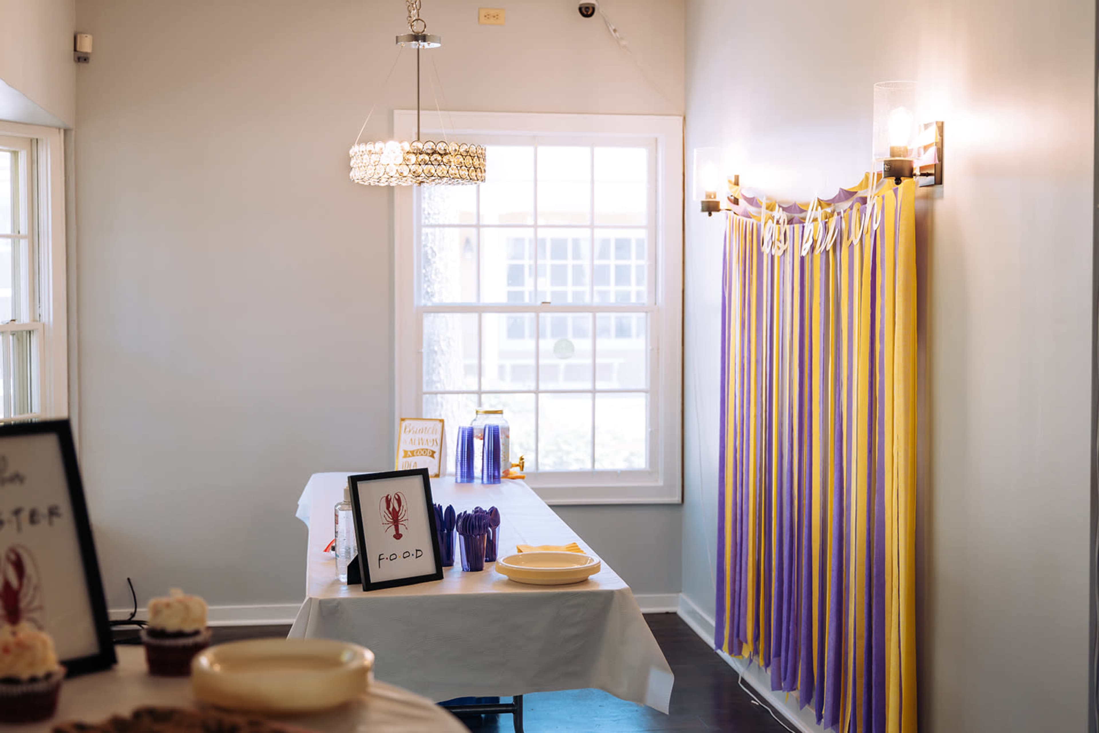 A decorated room with a table set for a gathering, featuring a white tablecloth, plates, and signs, alongside colorful streamers on the wall.
