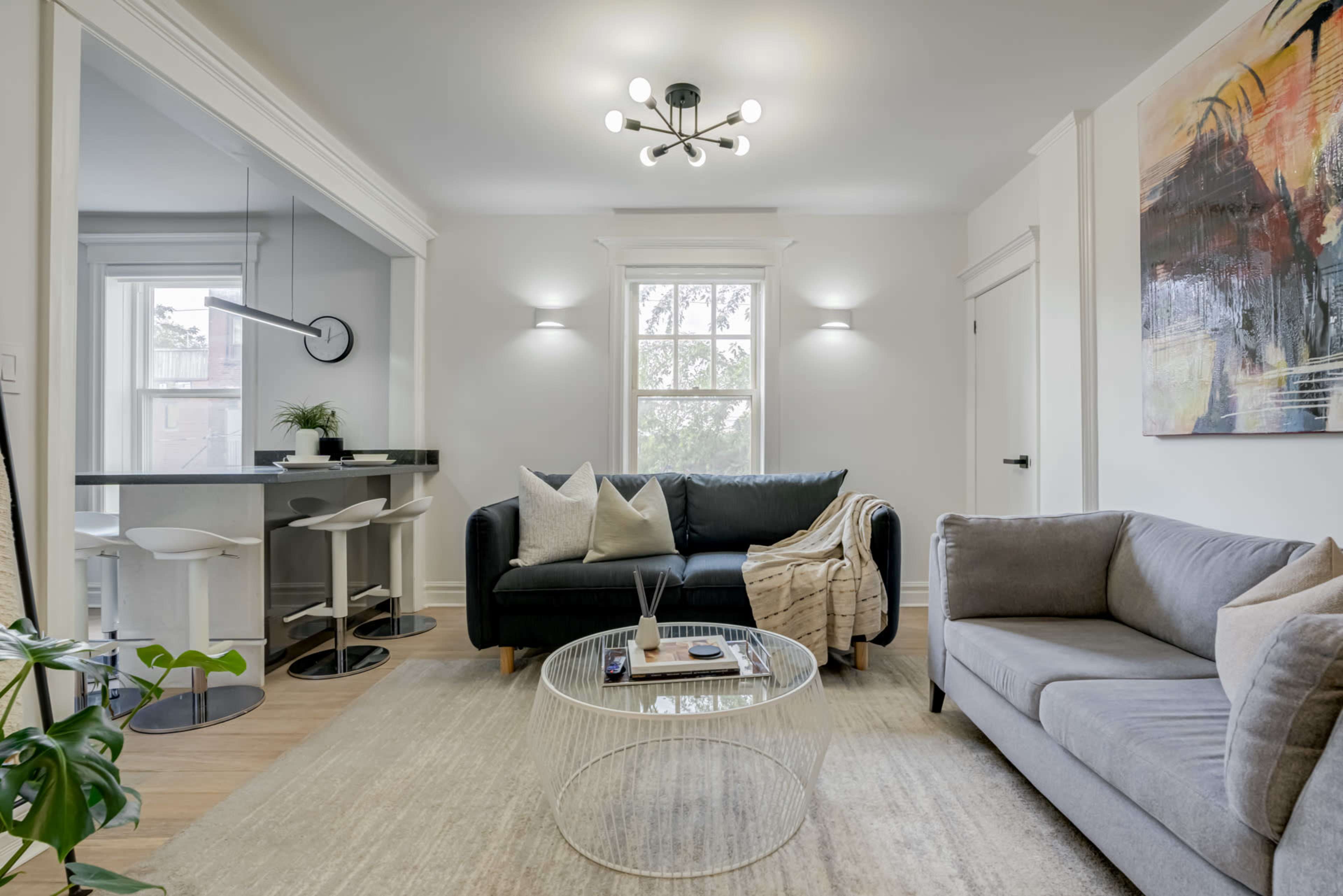 A modern living room with a black sofa, a gray sofa, a round glass coffee table, and a dining area in the background.