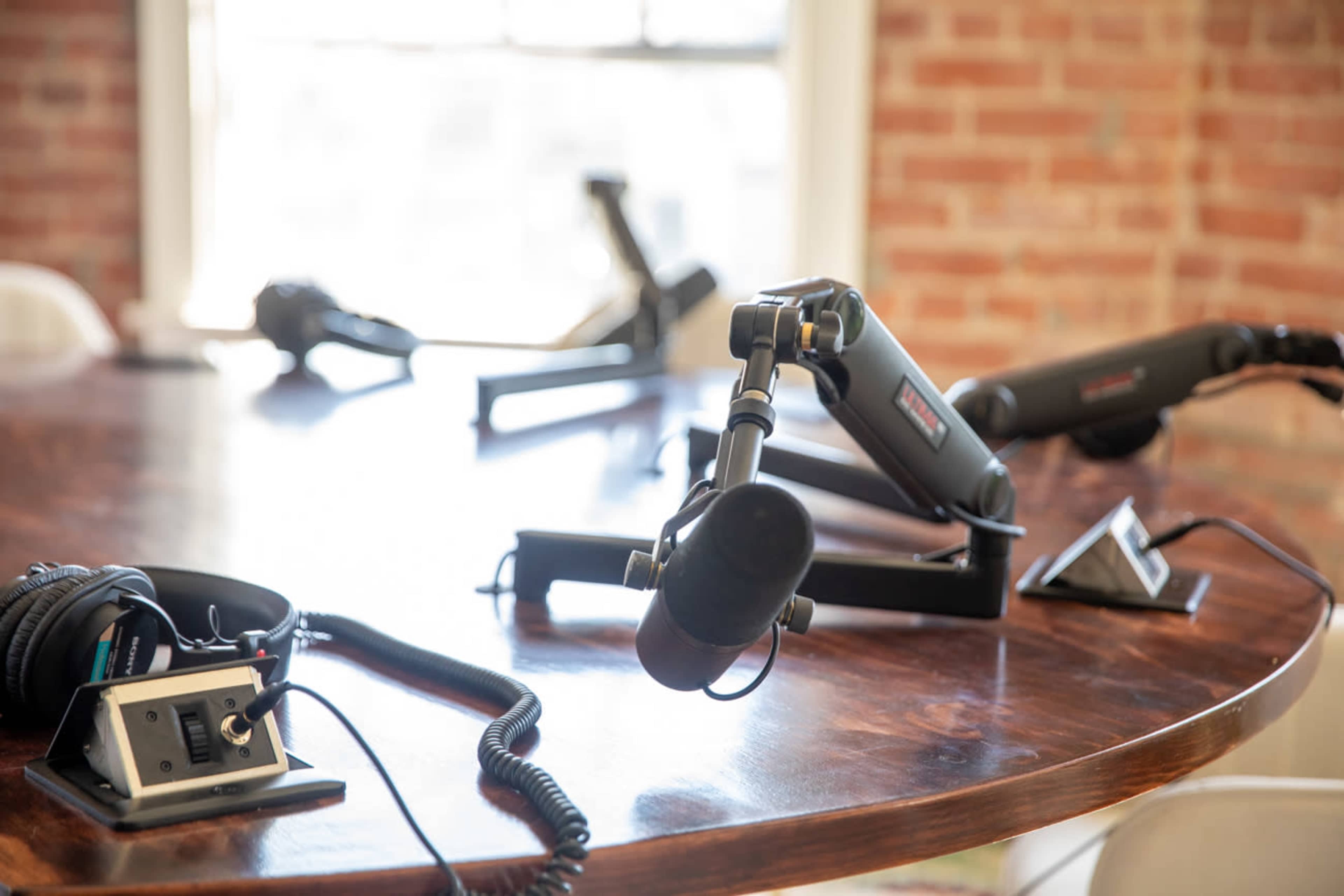 A table with mounted microphones, headphones, and audio equipment is set up in a room with exposed brick walls.