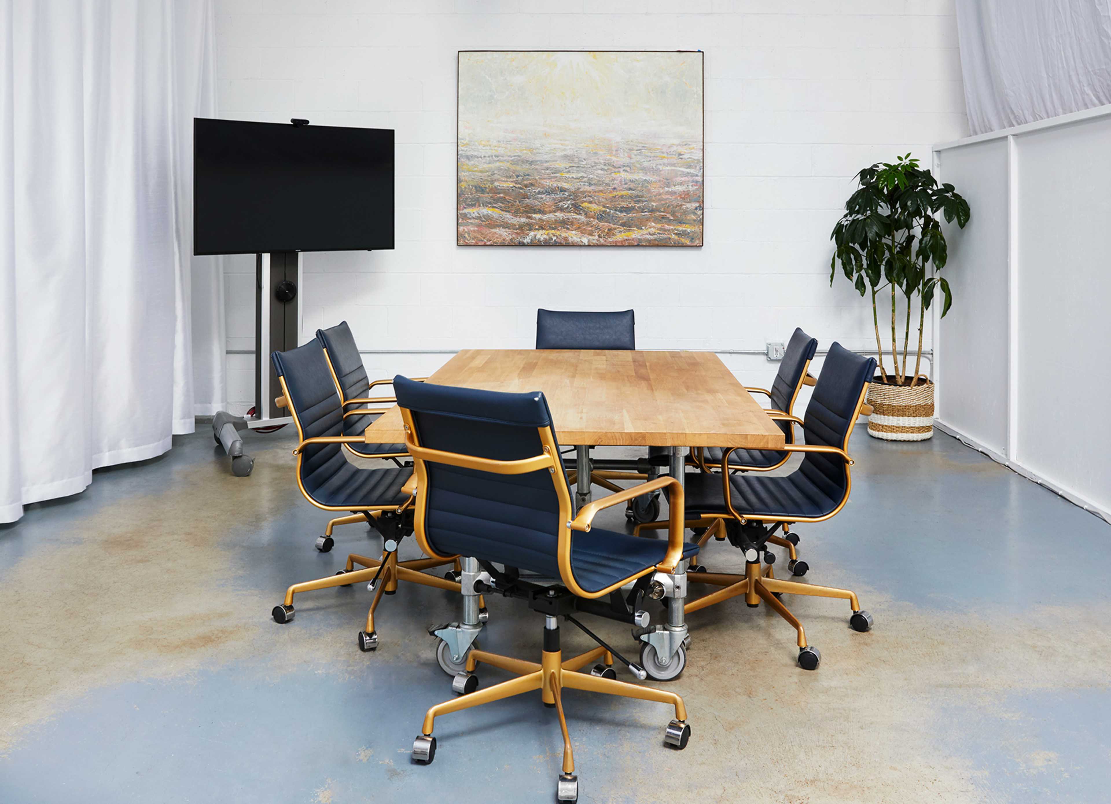 A modern conference room features a large wooden table surrounded by ergonomic chairs, with a television and a potted plant in the background.
