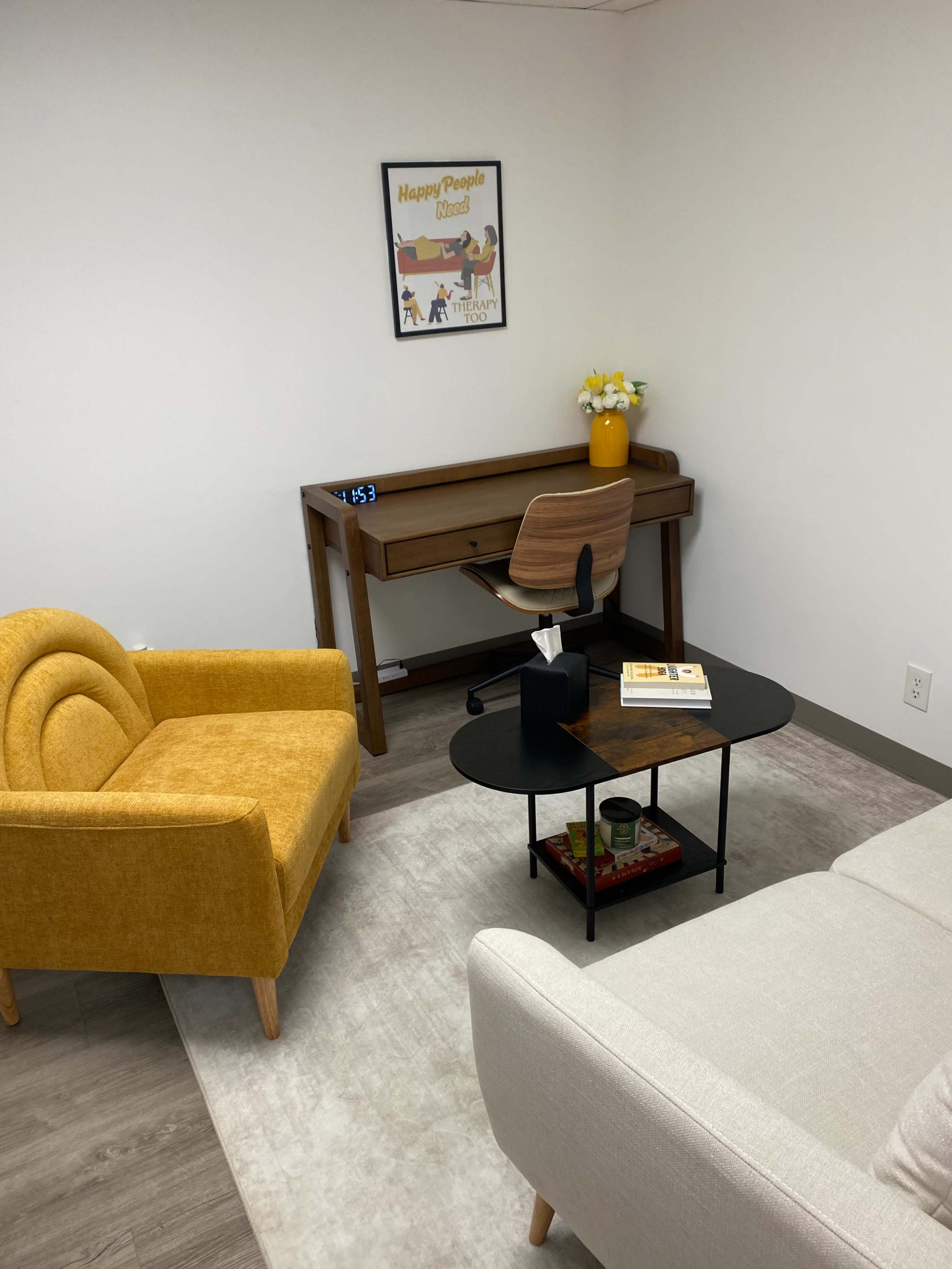 The image shows a small, organized office space featuring a yellow upholstered chair, a wooden desk with a clock and flowers, a circular black coffee table, and a light-colored sofa.
