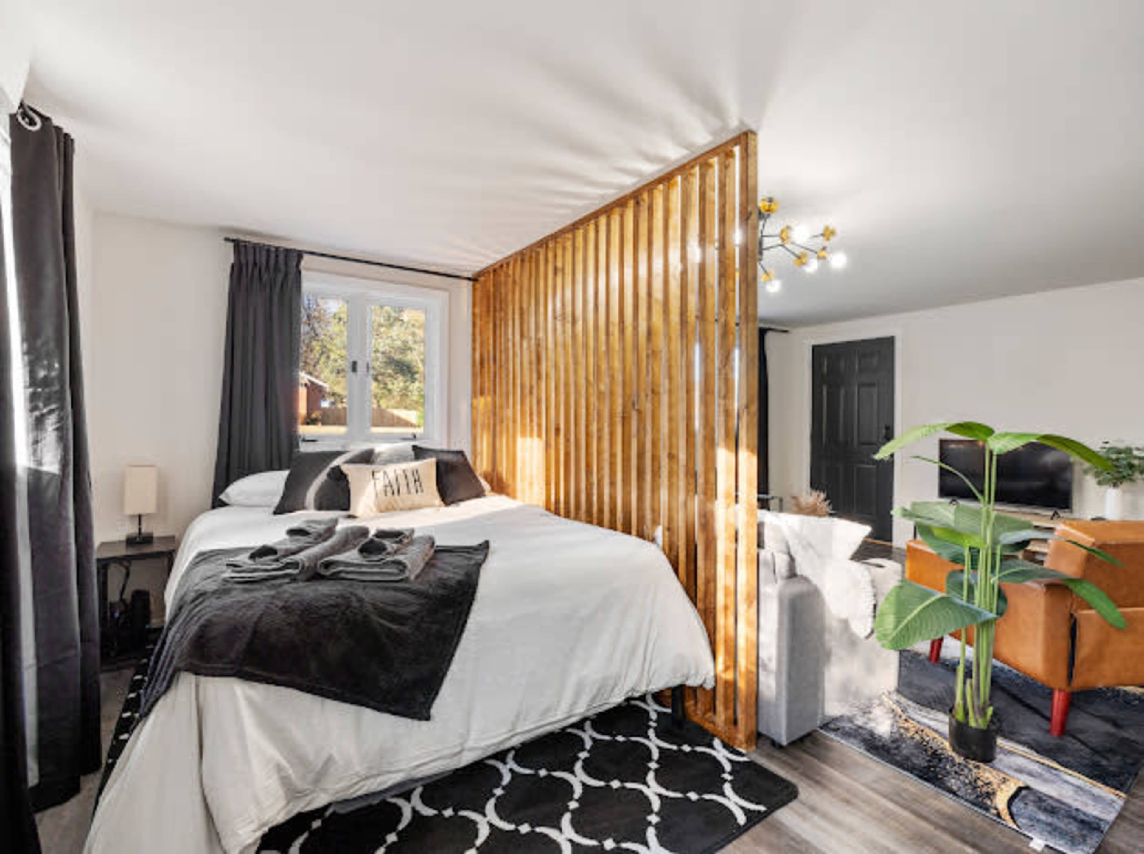 A modern bedroom with a wooden partition separating the sleeping area from a small living space, featuring a bed, couch, and potted plant.
