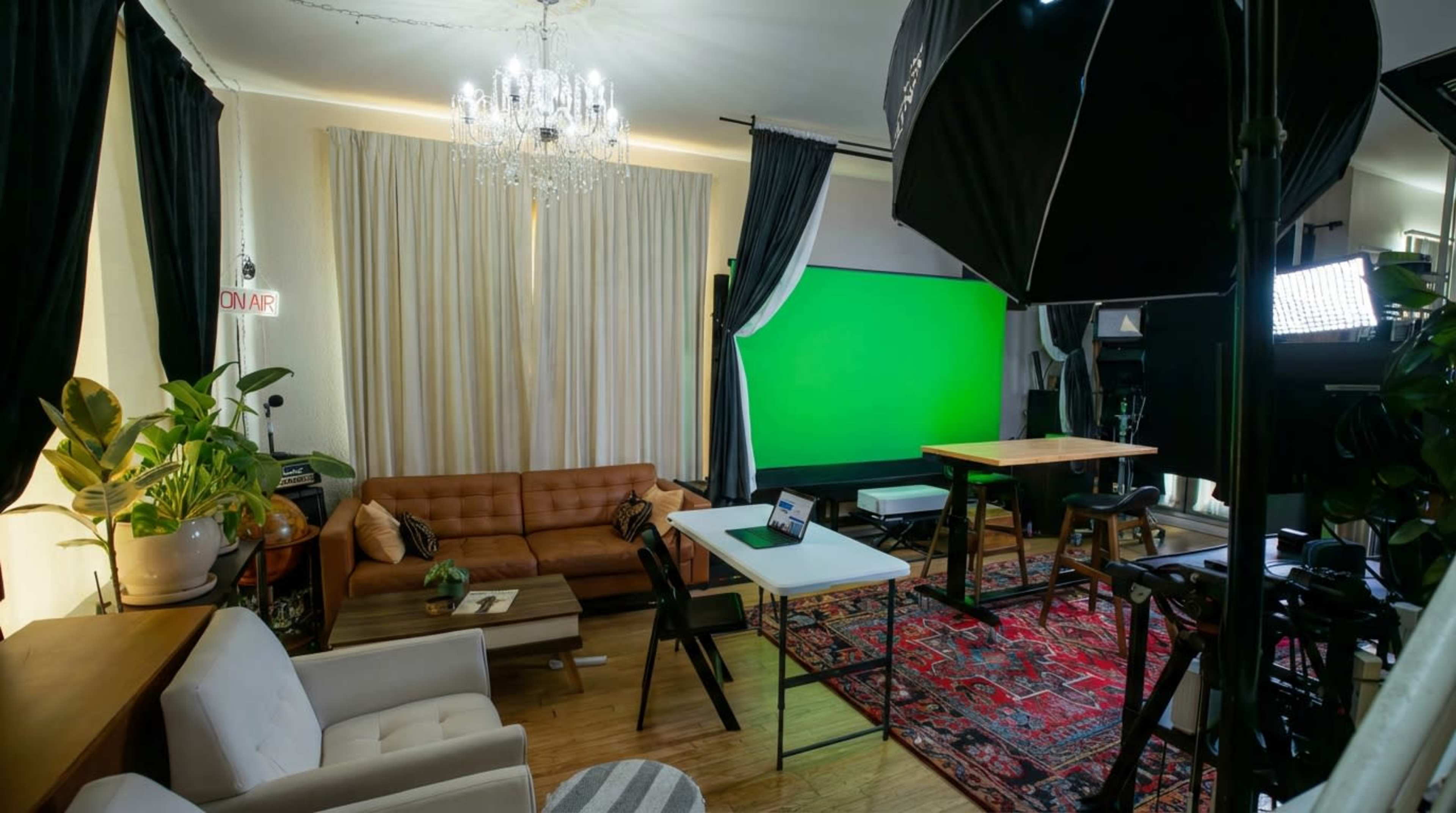 The image shows a well-lit studio space with a green screen, a brown leather couch, a wooden table, and various lighting equipment.