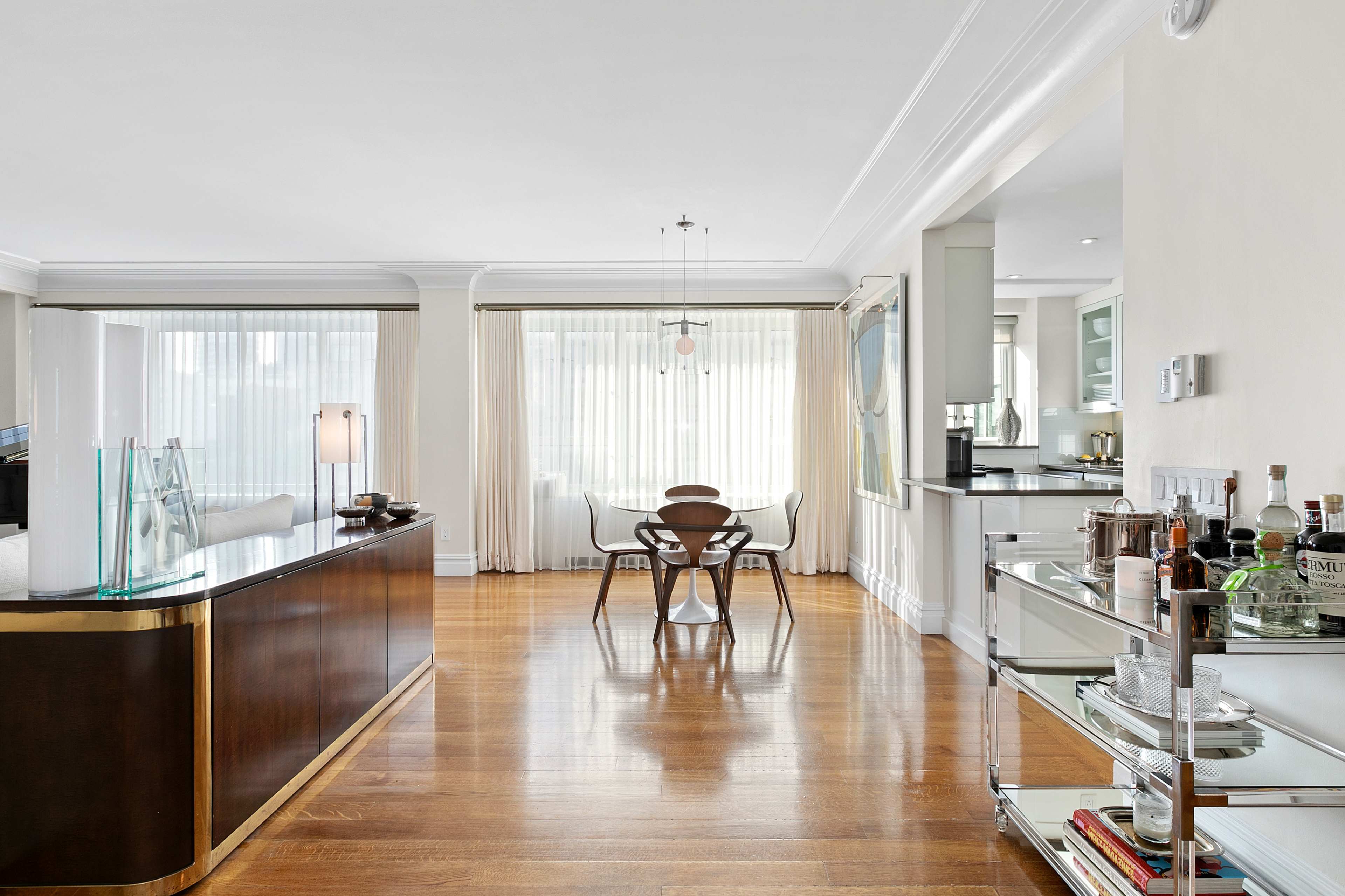 A modern kitchen and dining area with wooden flooring, large windows covered by sheer curtains, and a sleek bar cart.