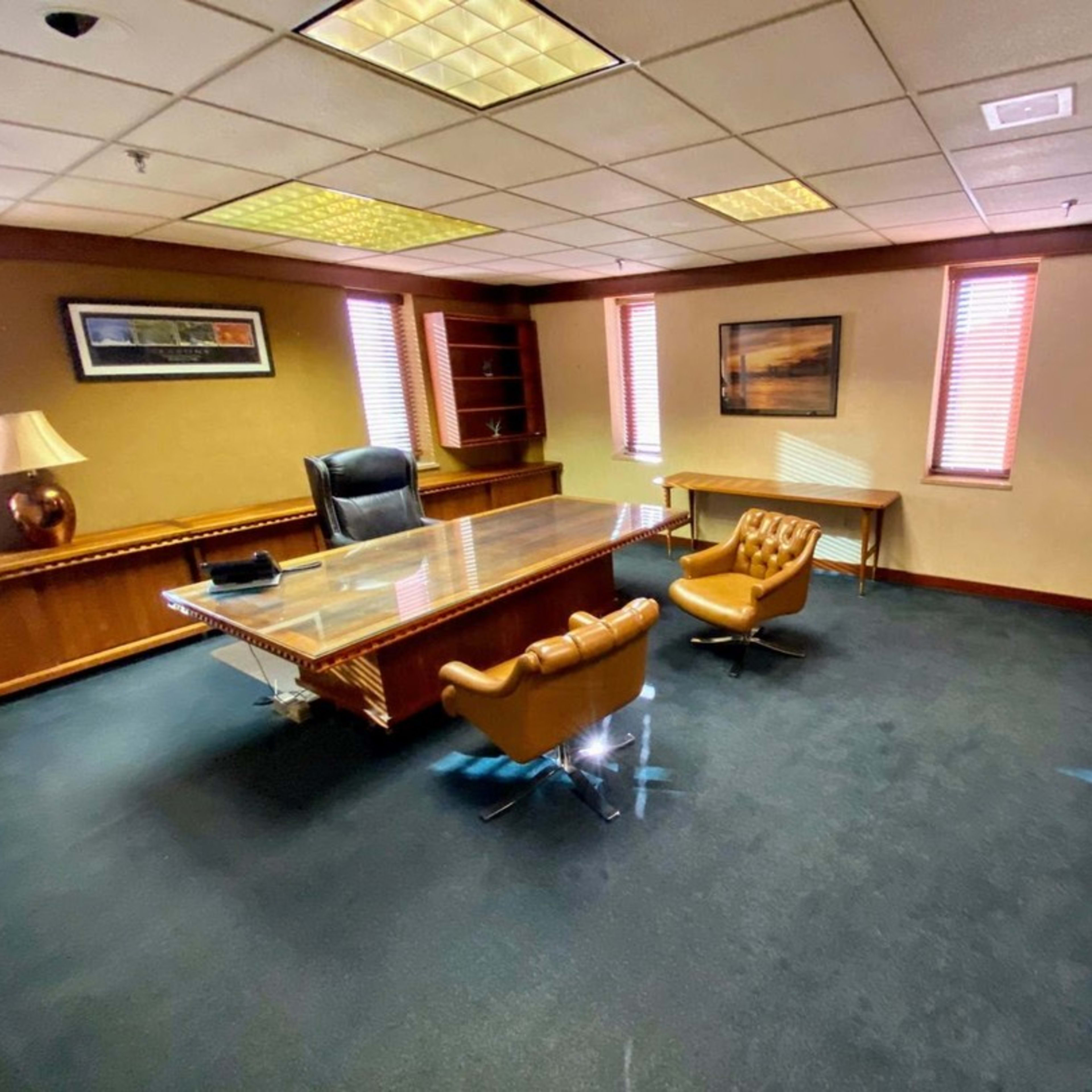 The image shows a large office with a wooden desk, two brown leather chairs, a bookshelf, and windows with blinds.