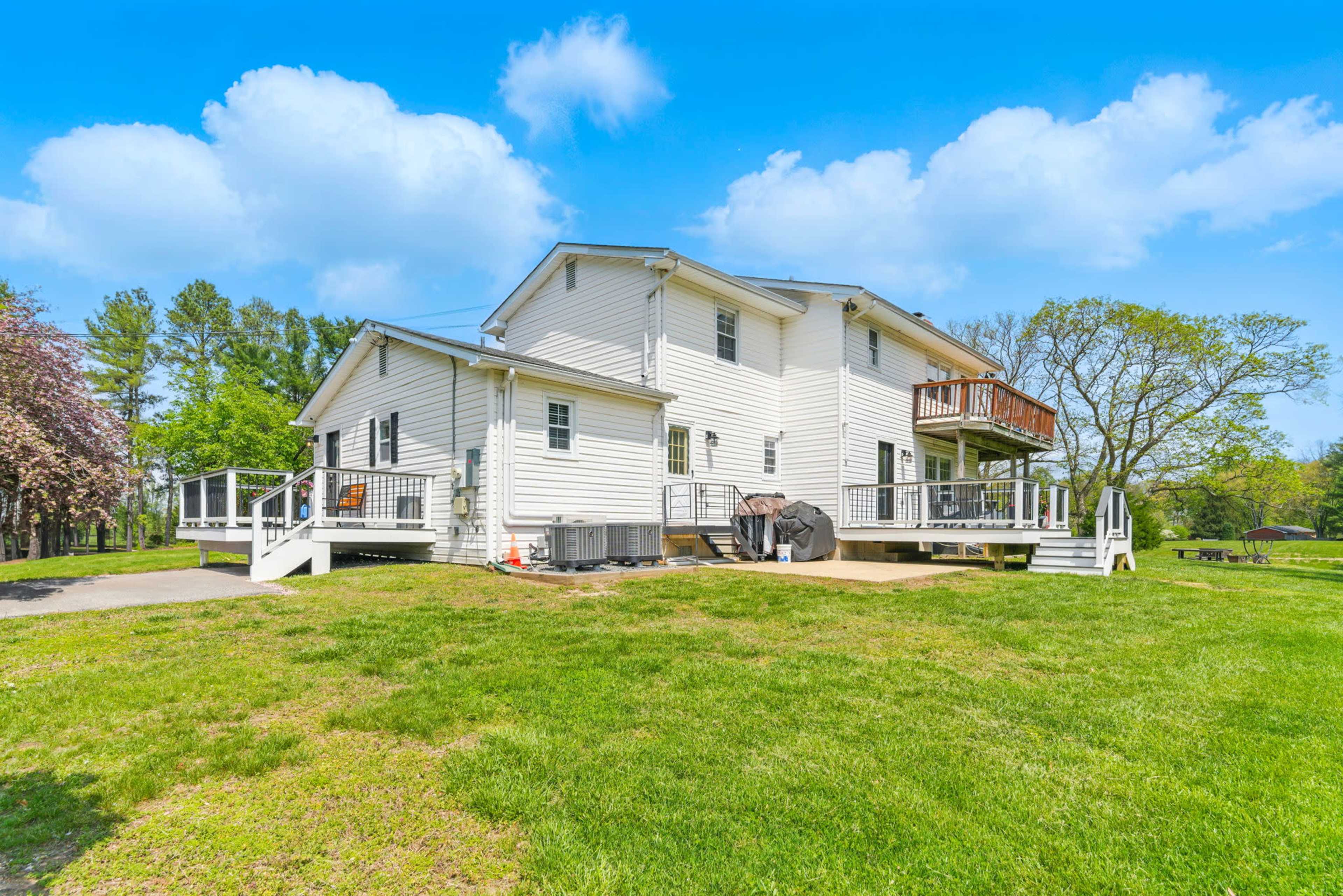 A two-story white house with a deck, surrounded by green grass and trees, sits against a clear blue sky.