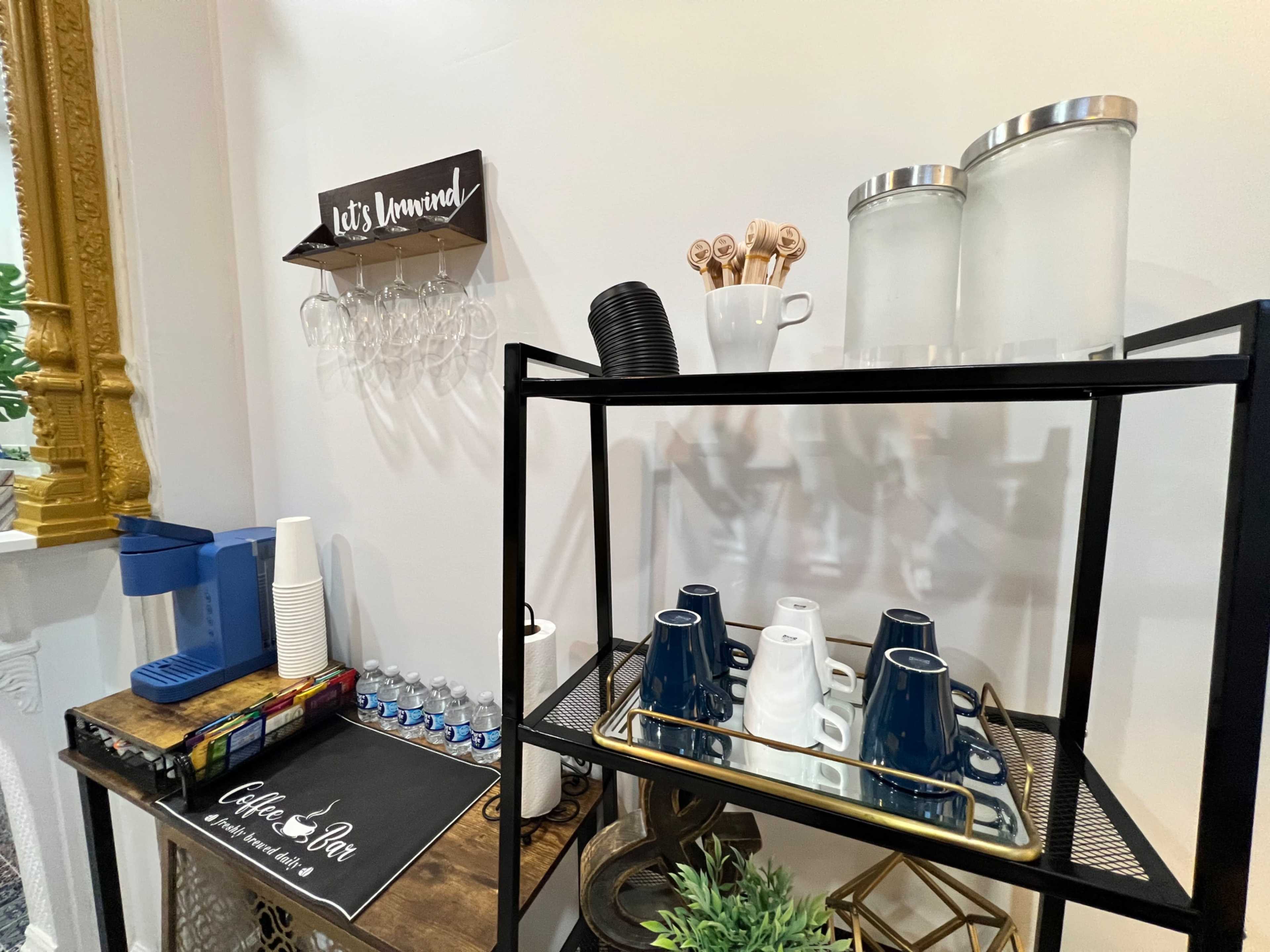 A coffee station features a variety of cups, water bottles, and utensils, with a decorative sign above.