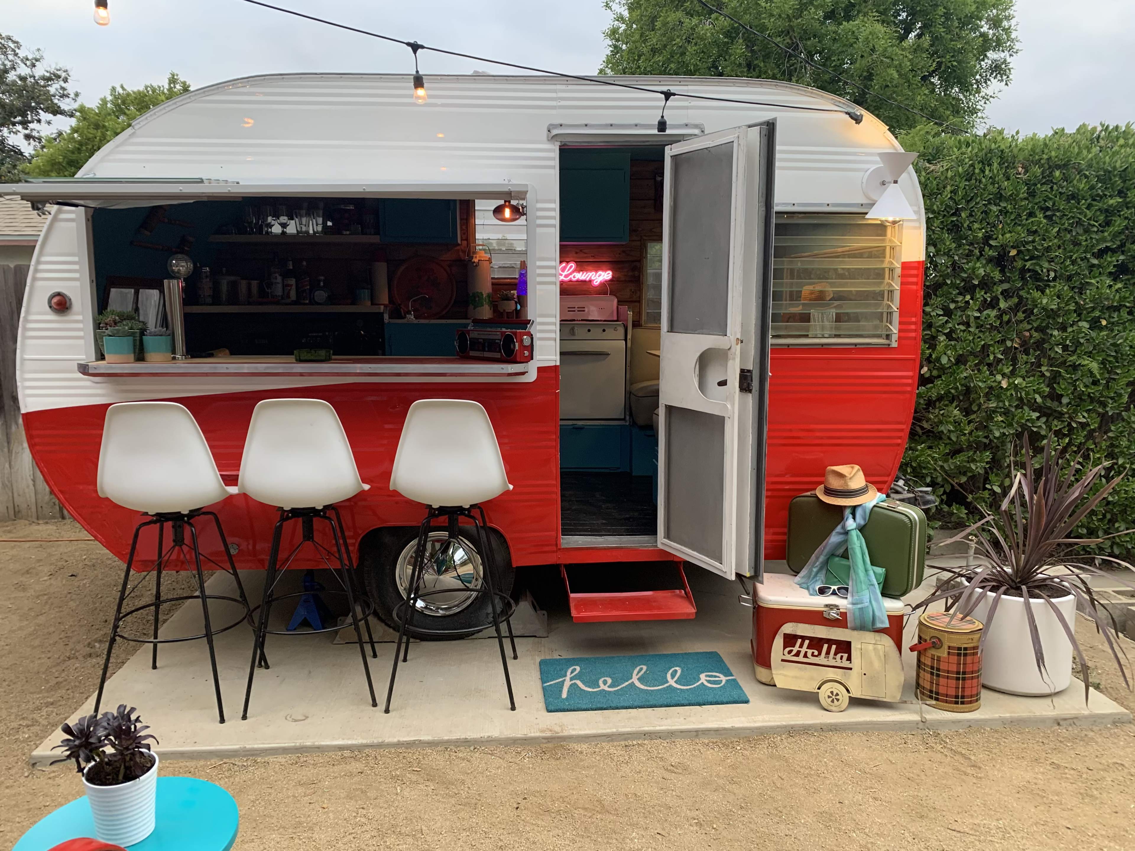 A vintage red and white camper trailer features an open door with bar stools outside, accompanied by various decorative items and plants nearby.