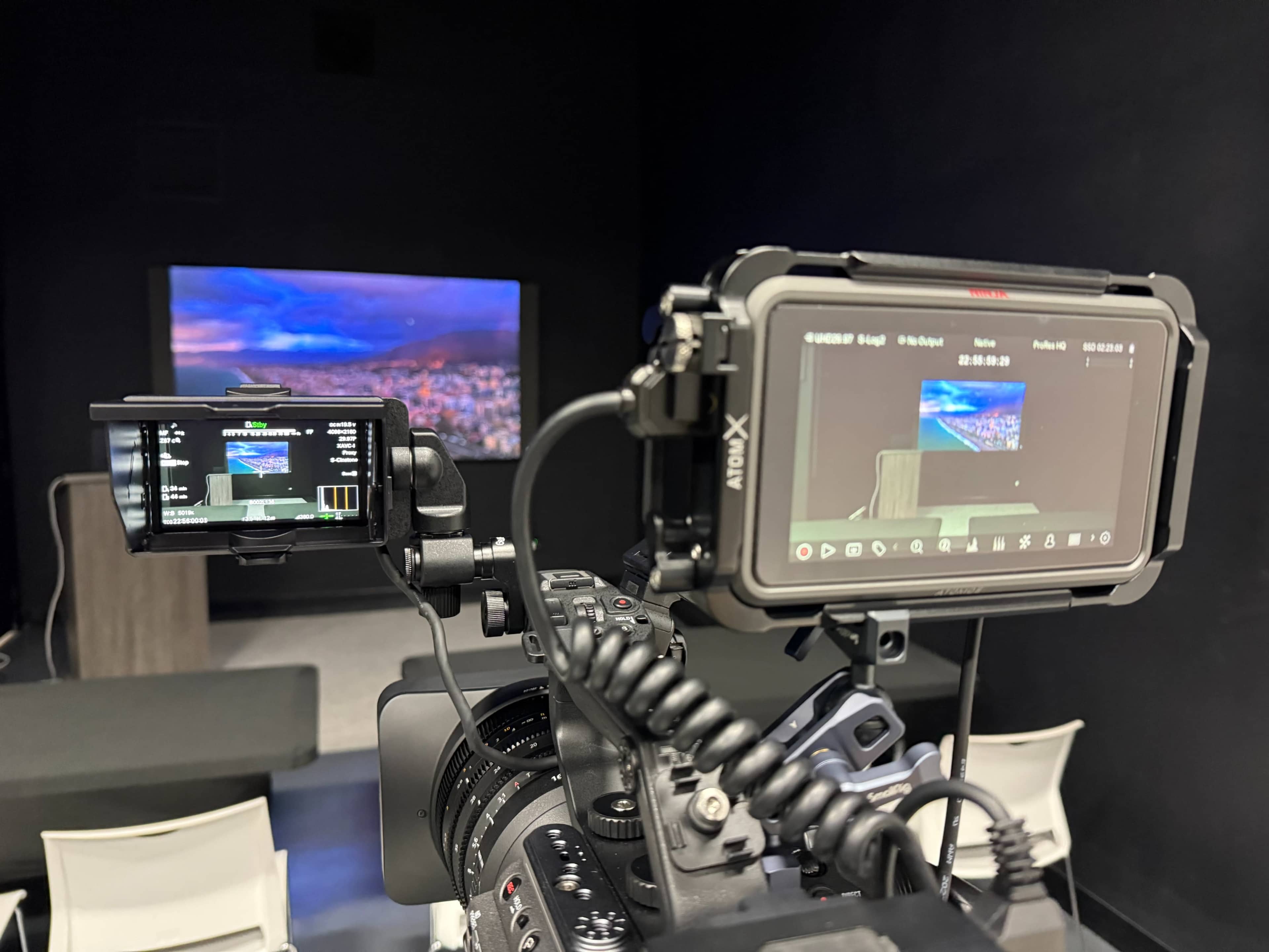 The image shows two video cameras positioned on a tripod, with screens displaying a presentation on a large monitor in a dark room.