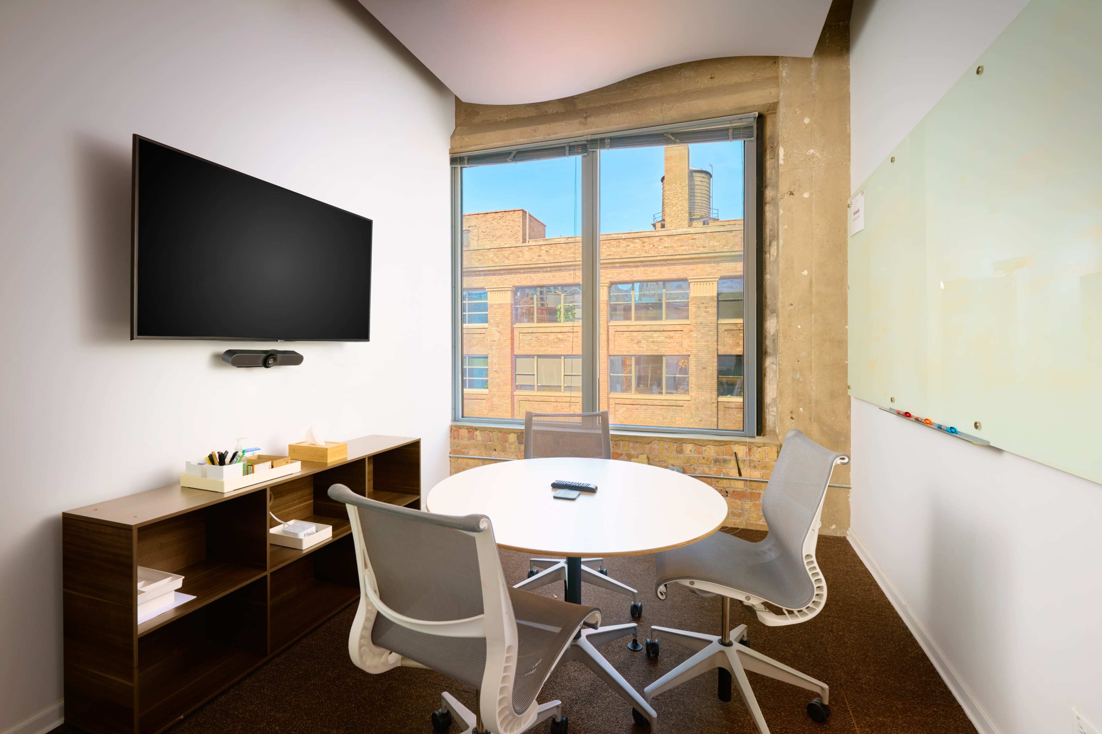 A small conference room features a round table surrounded by four chairs, a wall-mounted TV, and a large window overlooking a brick building.