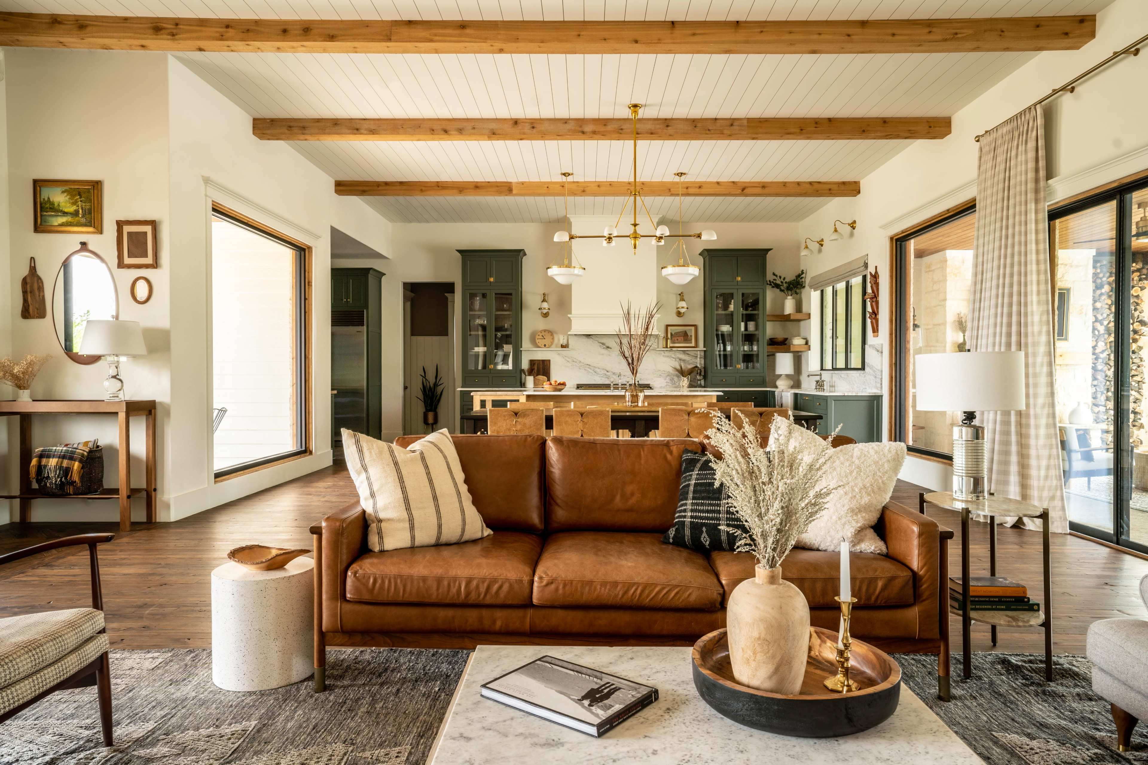 A modern living room featuring a brown leather sofa, a marble coffee table, and large windows that lead to a kitchen with green cabinetry.