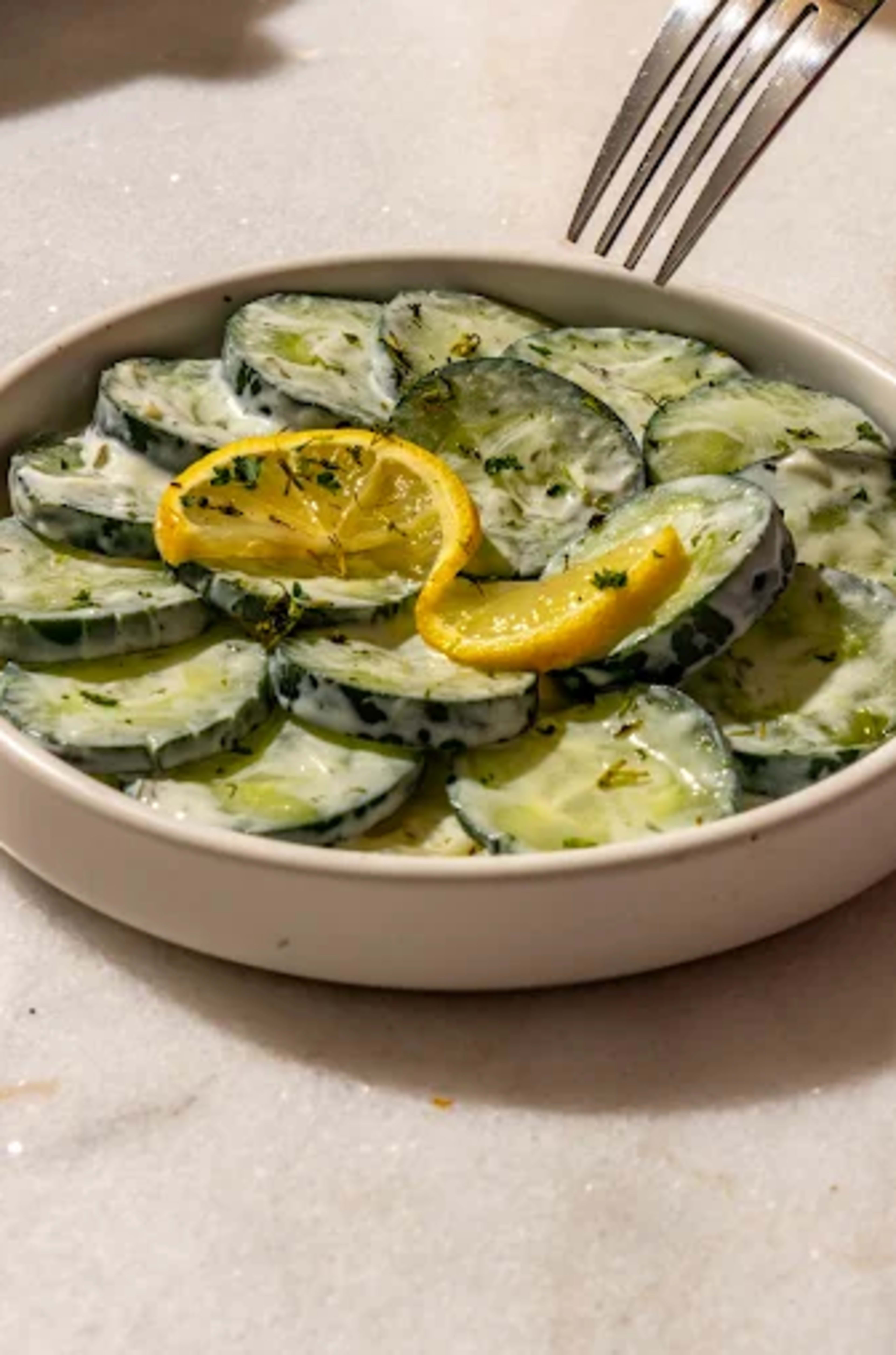 A white bowl contains sliced cucumbers topped with lemon slices and sprinkled with herbs.