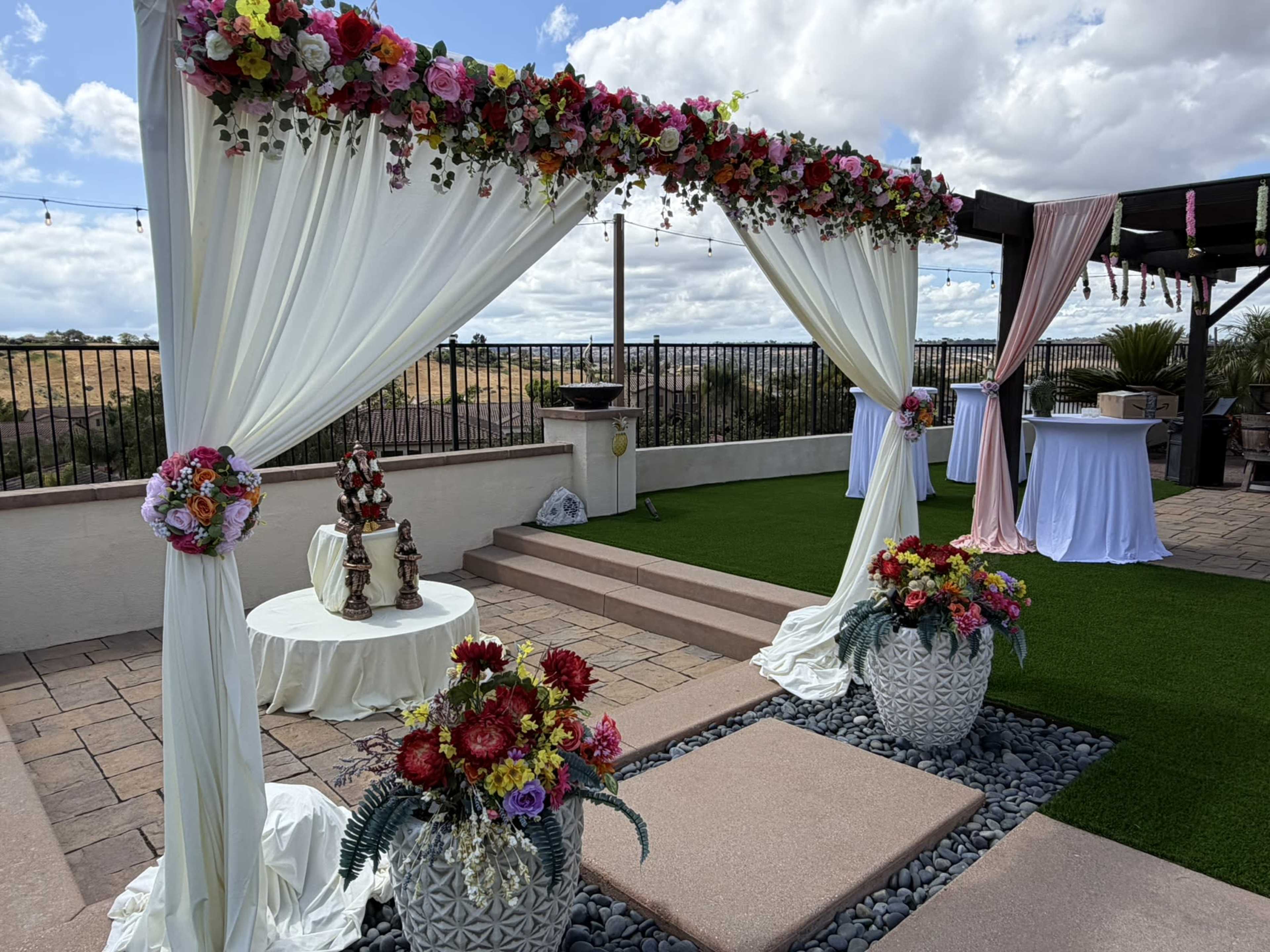 The image shows a decorated outdoor event area with floral arrangements on a white draped arch, surrounded by potted flowers, stone flooring, and tables set up in the background.