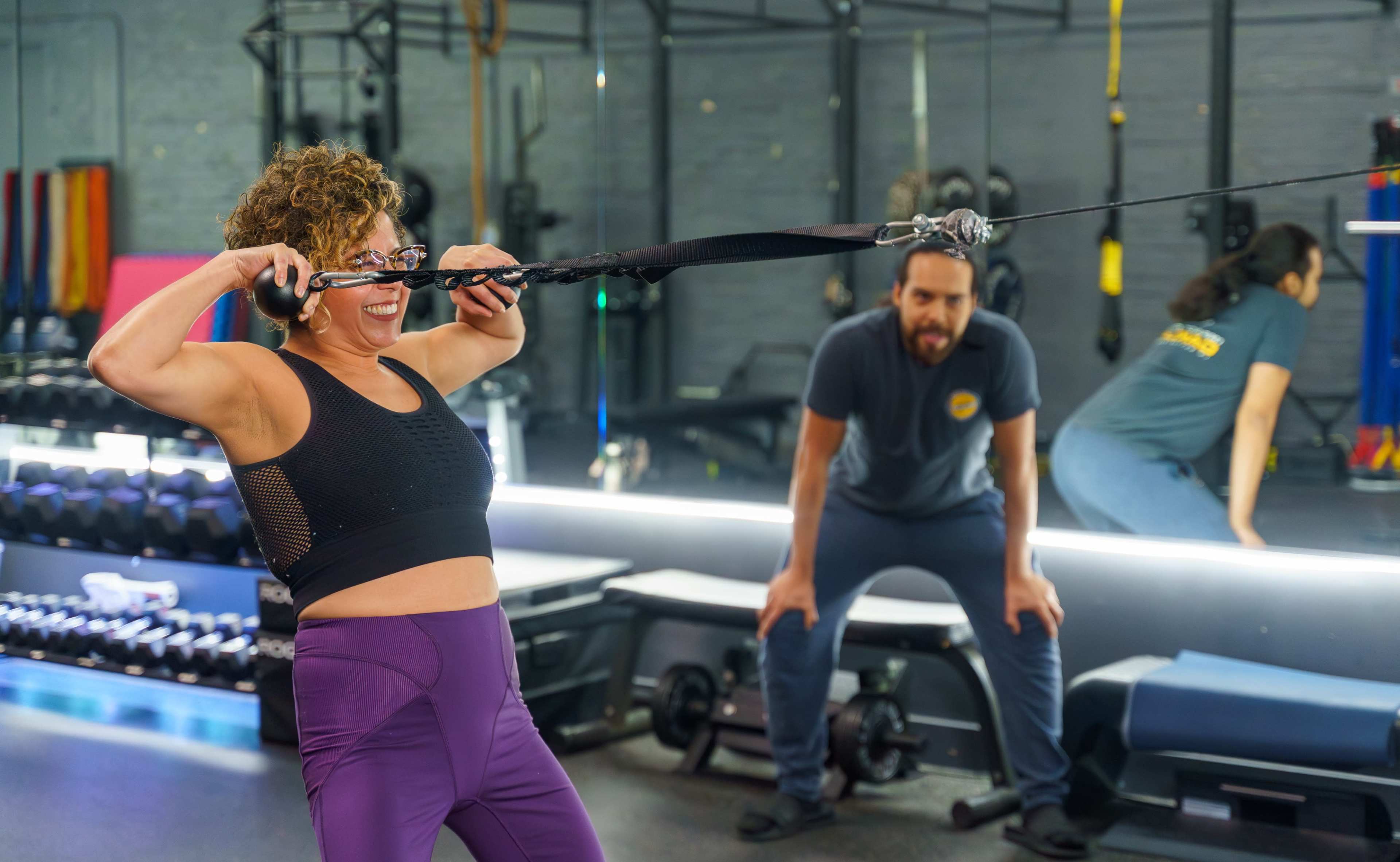 A woman performs a cable exercise in a gym while a man coaches her from behind.