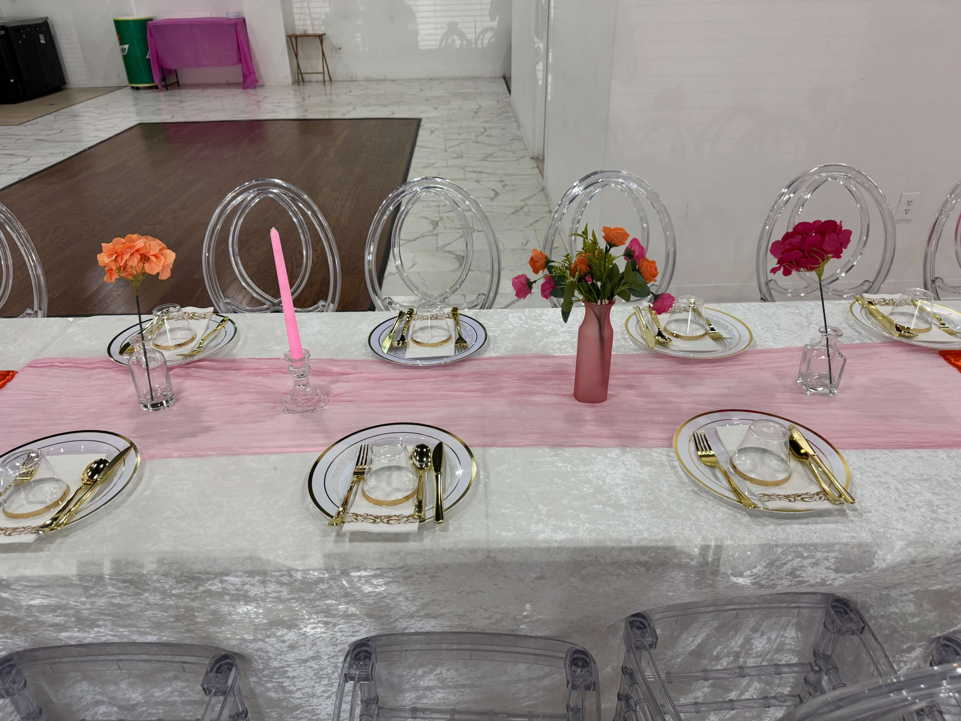 A long dining table is set with clear acrylic chairs, white tablecloth, plates, utensils, and a central pink runner adorned with flowers in small vases.