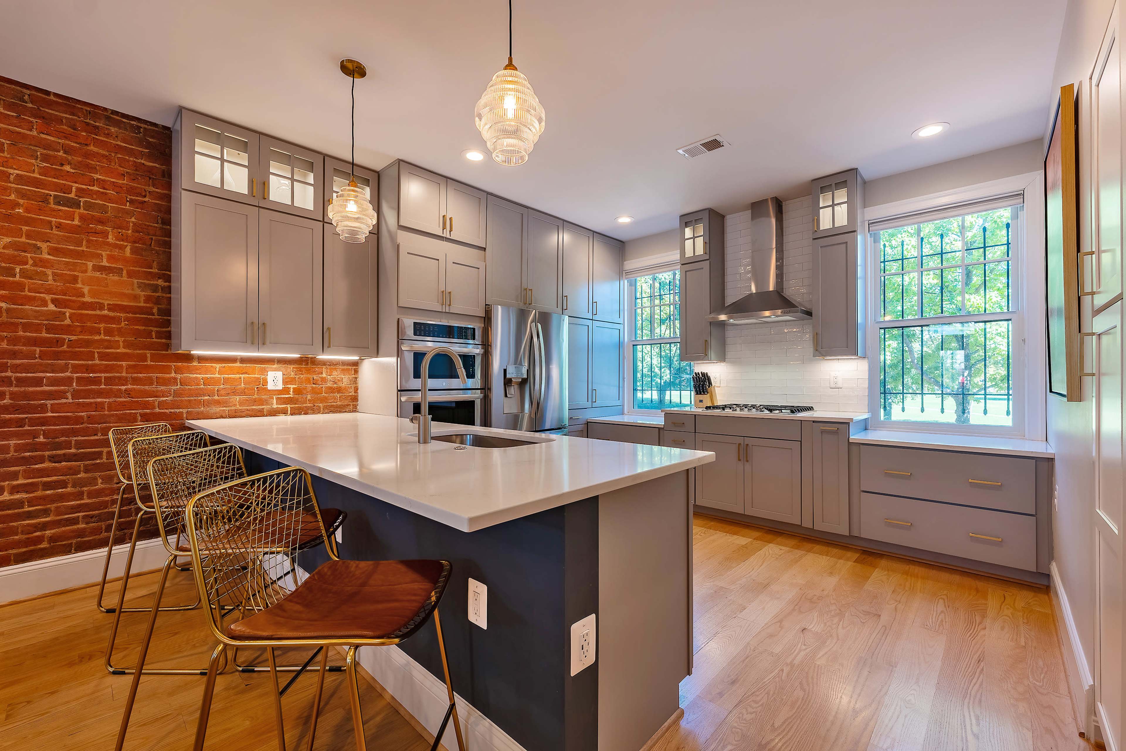 A modern kitchen with gray cabinetry, a white countertop, a brick accent wall, and pendant lighting over a breakfast bar with metal stools.