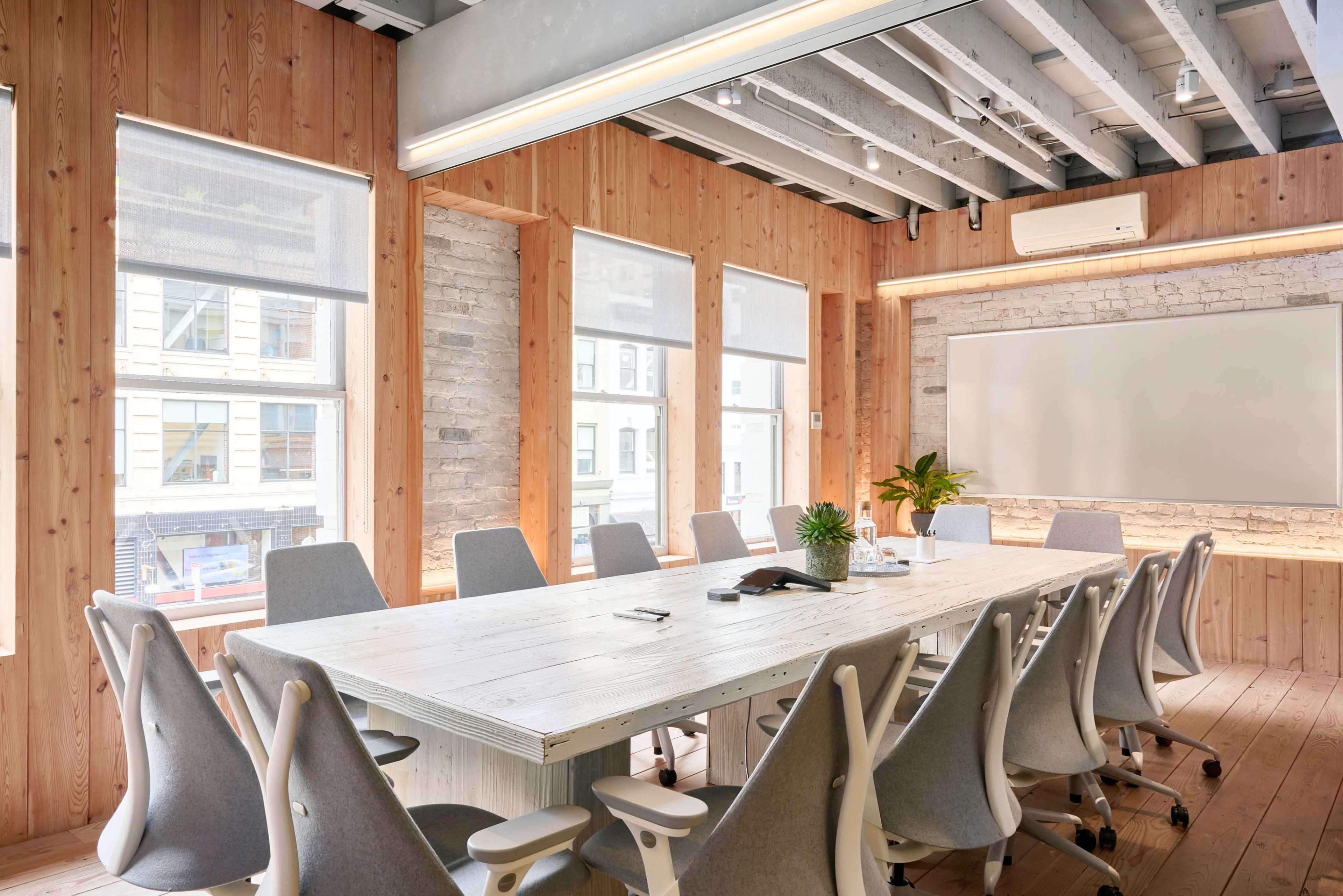 The image shows a modern conference room with a long wooden table surrounded by ergonomic chairs and large windows providing natural light.