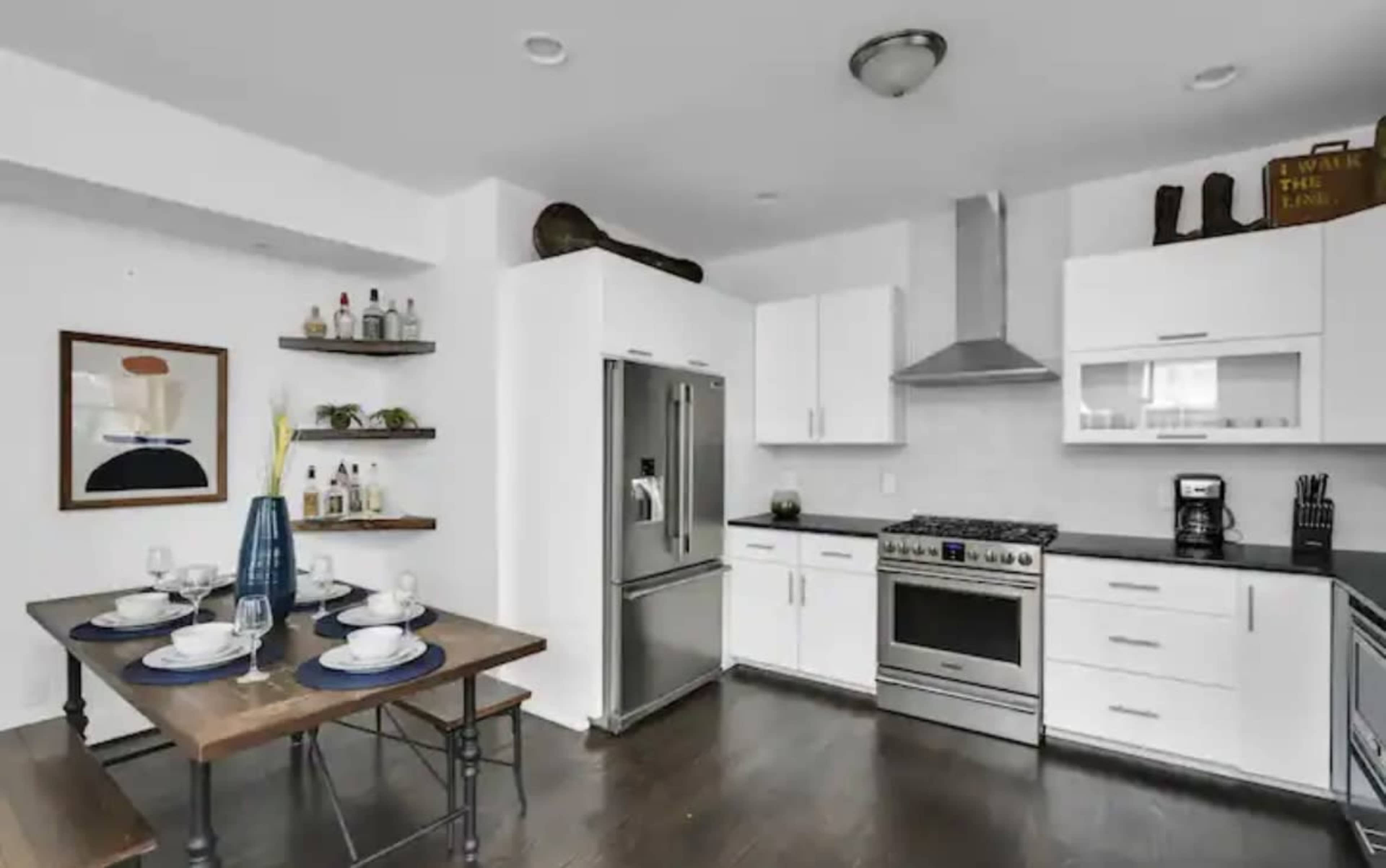 A modern kitchen with white cabinetry, stainless steel appliances, a wooden dining table set for four, and open shelving displaying various bottles and decor items.